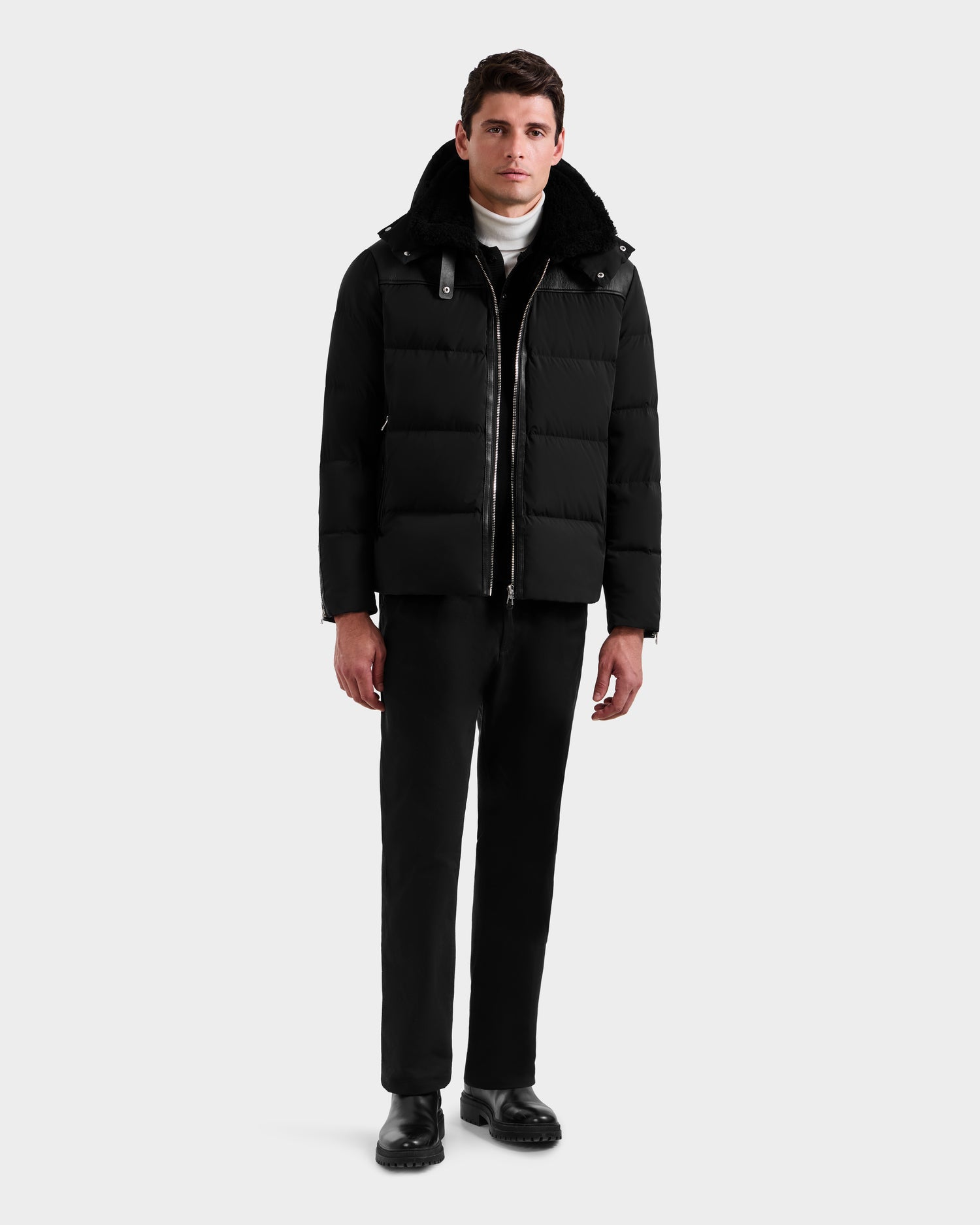 A man faces forward wearing the Rudsak SIMON - 6125597 Black men’s quilted down puffer with a shearling combo collar, black pants, black shoes, and a white turtleneck sweater against a plain light gray background.
