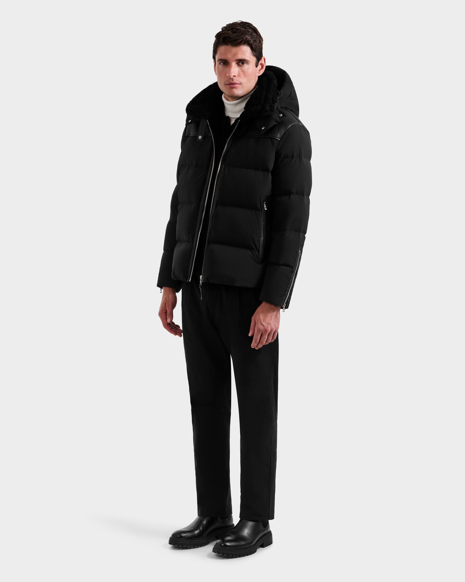 A man stands against a plain background wearing the Rudsak SIMON - 6125597 Black men’s down puffer jacket with a hood, paired with black pants, black boots, and a white turtleneck.