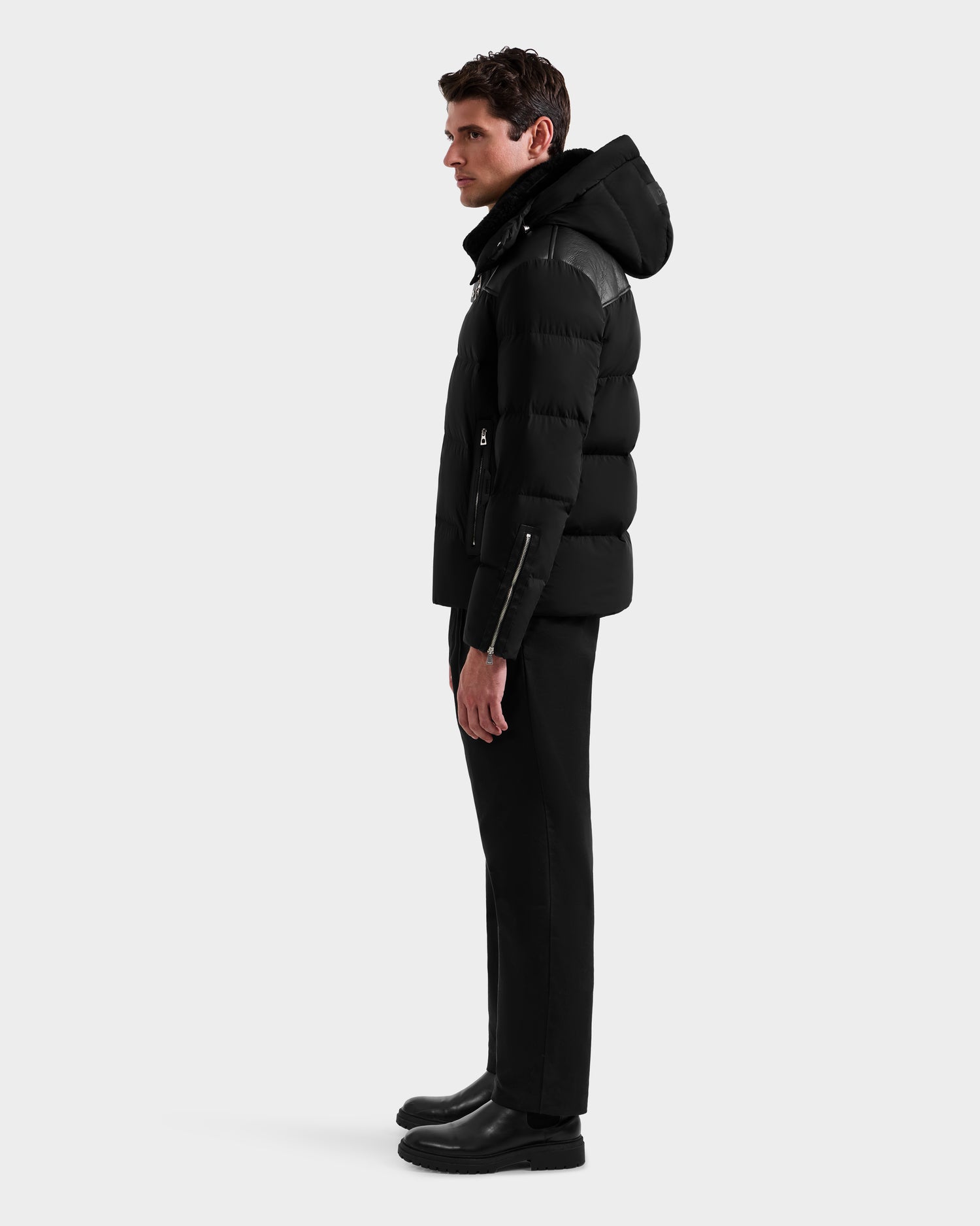 A man stands in profile against a plain background, wearing the Rudsak SIMON - 6125597 Black men’s down puffer winter jacket with a hood, paired with black pants and black boots.