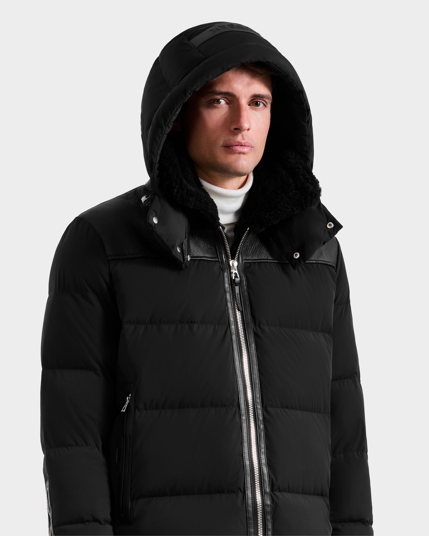 A man wearing the Rudsak SIMON - 6125597 Black men’s down puffer jacket with a hood and a white turtleneck stands against a plain light gray background, looking at the camera with a neutral expression.