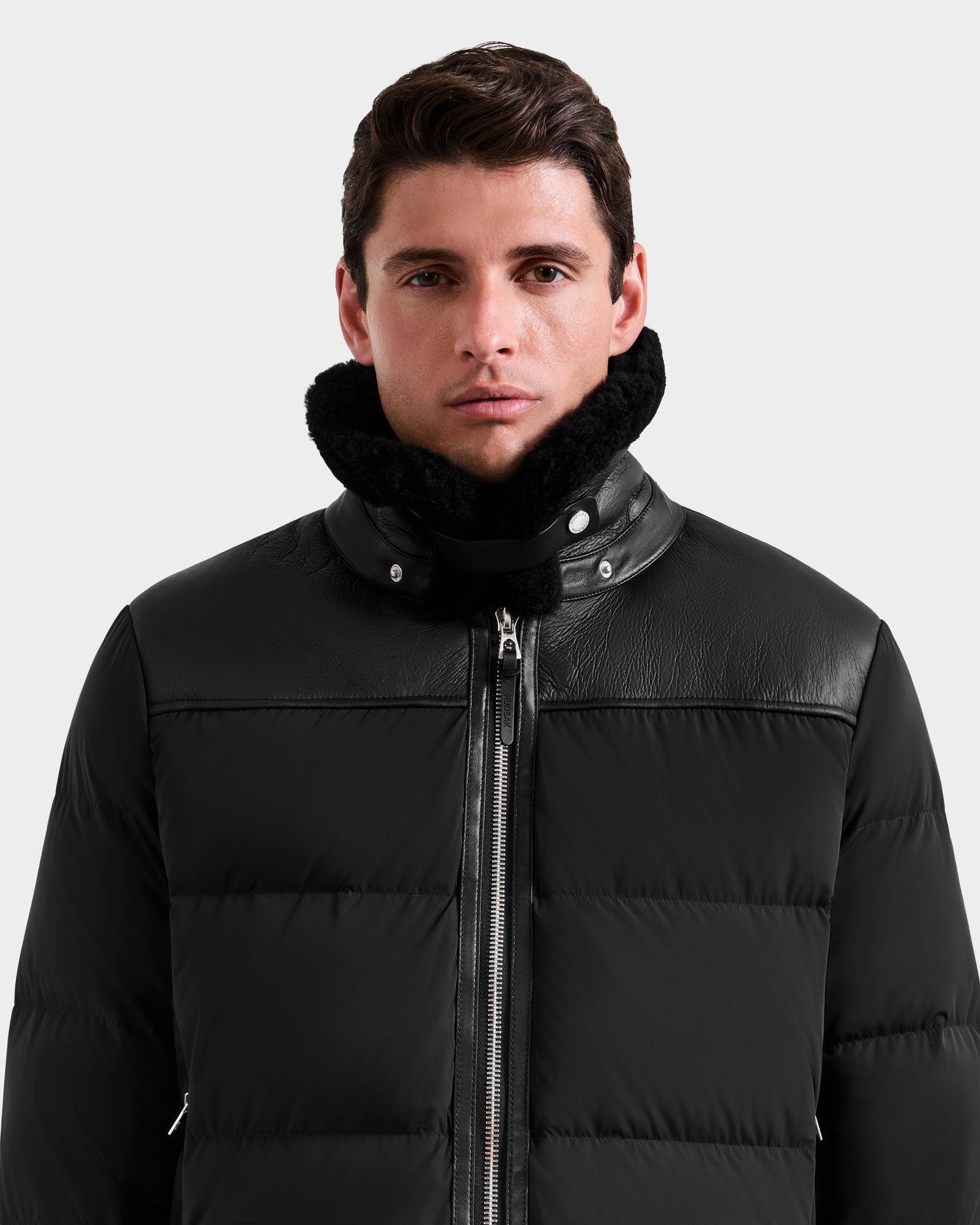 A man with short dark hair models the Rudsak SIMON - 6125597 Black men's quilted down puffer jacket with a high shearling collar, standing against a plain light gray background.