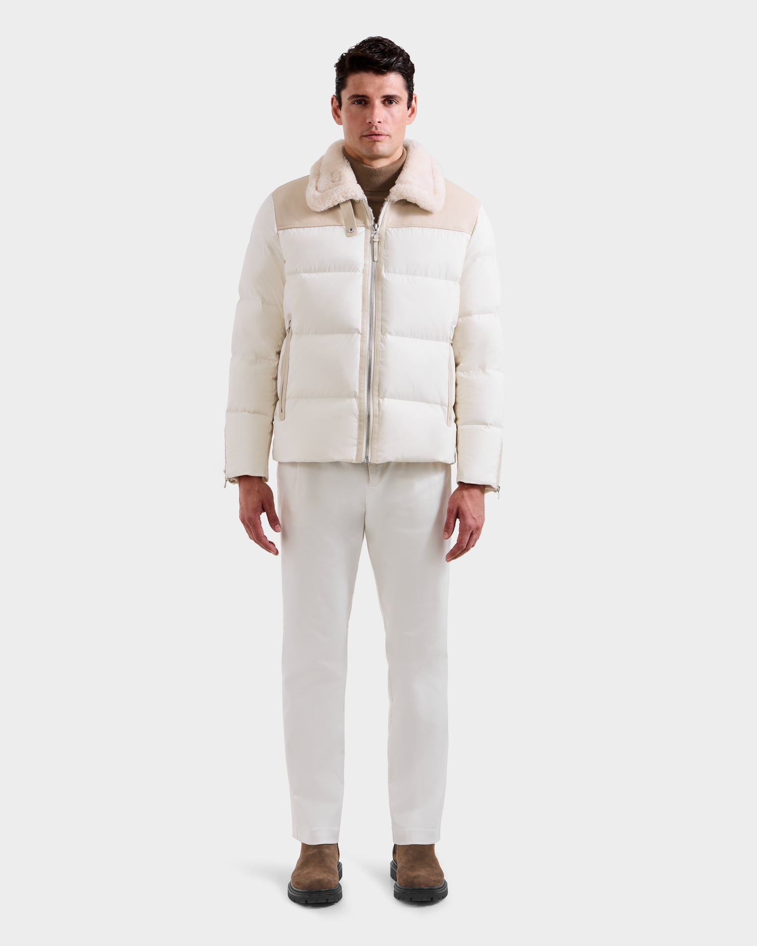 A man faces forward wearing the SIMON - 6125597 Vanilla men’s down puffer with shearling collar by Rudsak, paired with white pants, a beige shirt, and brown shoes, set against a plain white background.
