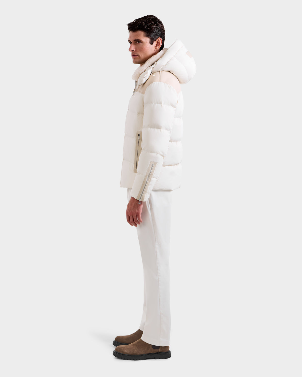 A man in profile wears the Rudsak SIMON - 6125597 Vanilla quilted down puffer jacket with a hood, paired with white pants and brown suede boots against a plain light background.