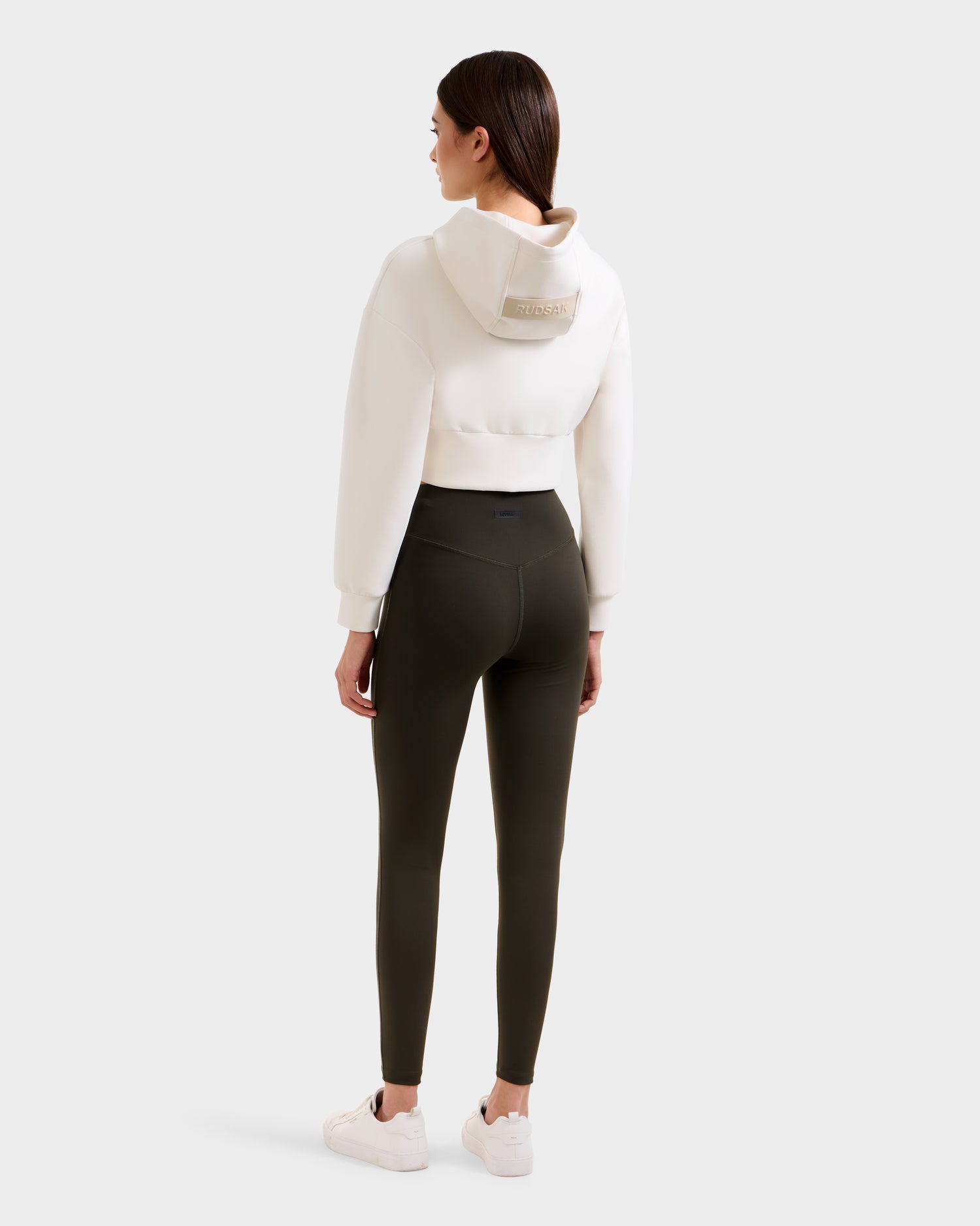 A woman with straight brown hair, seen from behind, wears a white cropped hoodie and Rudsak SONOMA - 8525071 Dark Olive leggings. She pairs the look with white sneakers against a plain light gray backdrop.