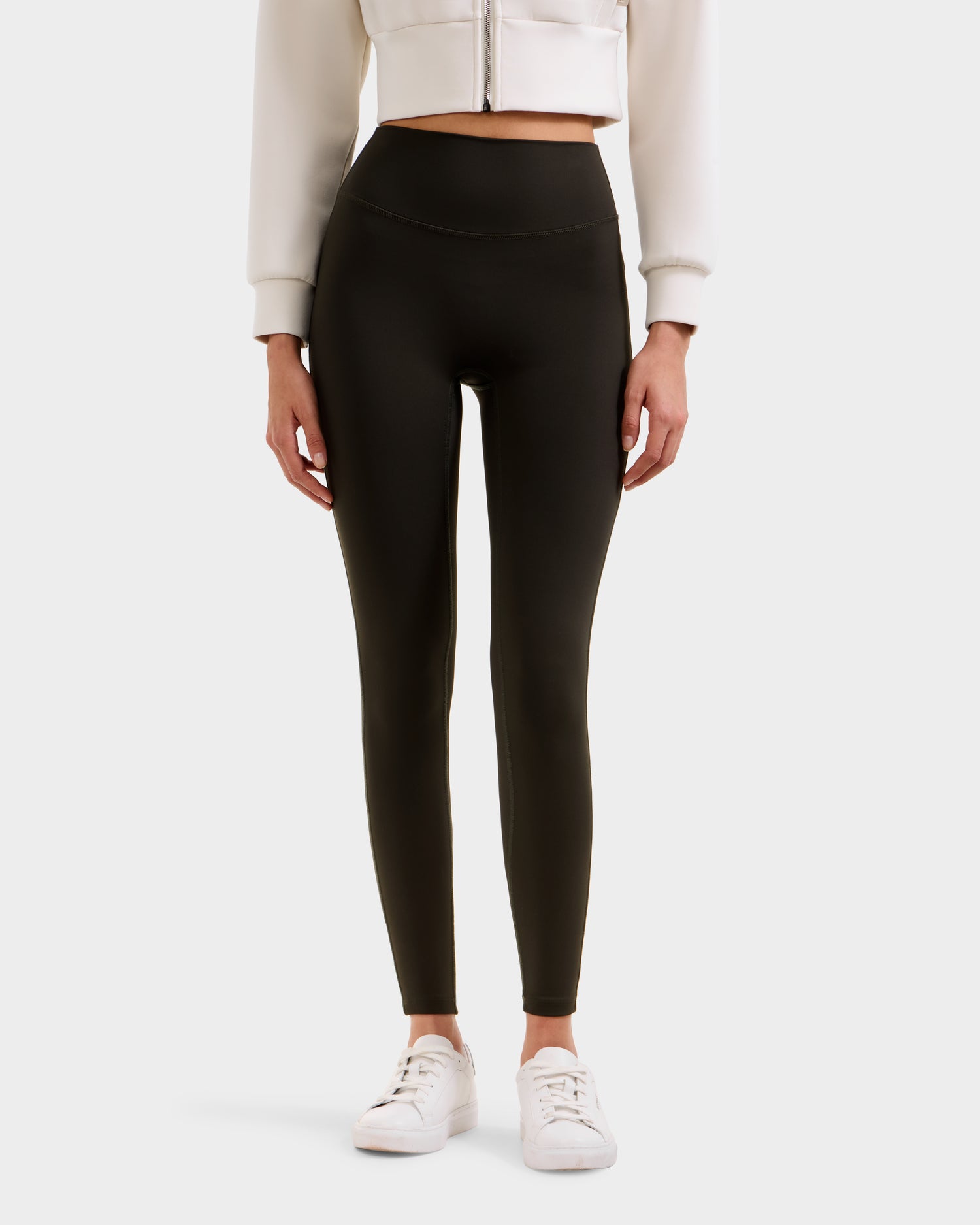 A person wearing high-waisted Rudsak SONOMA - 8525071 Dark Olive leggings, a white cropped jacket, and white sneakers stands against a plain background; only the body from shoulders down is visible.