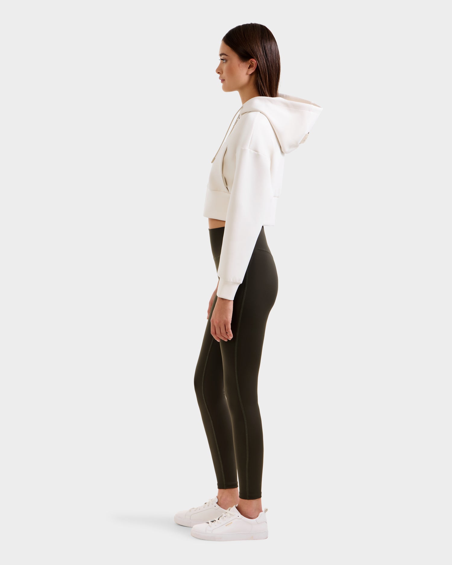 A woman stands in profile wearing a white cropped hoodie, Rudsak SONOMA - 8525071 Dark Olive leggings, and white sneakers against a plain light background.
