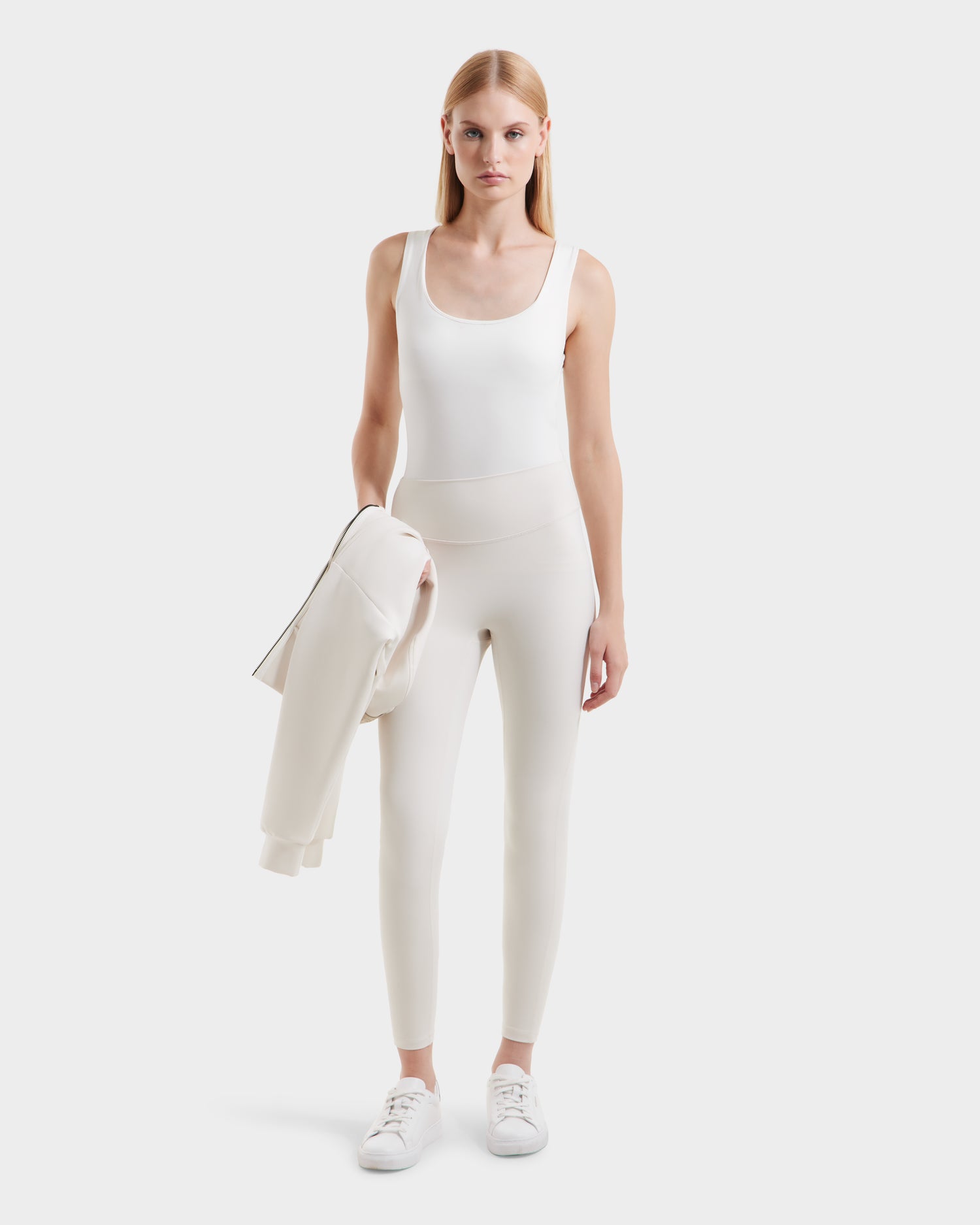A woman with straight blonde hair wears a white sleeveless top, high-waisted light beige Rudsak SONOMA - 8525071 Vanilla leggings, and white sneakers. She holds a light jacket in her left hand, standing against a plain white background.