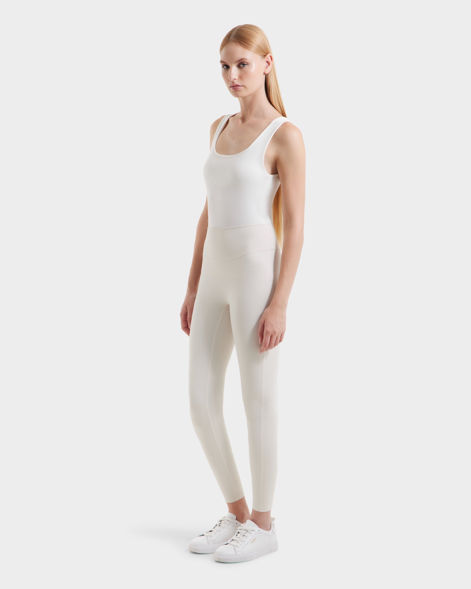 A woman with long blonde hair wearing a fitted white tank top, high-waisted Rudsak SONOMA - 8525071 Vanilla leggings, and white sneakers stands against a plain white background.