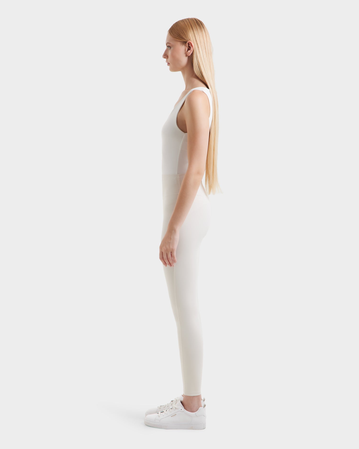 A woman with long straight blonde hair stands in profile, wearing a white sleeveless top, Rudsak SONOMA - 8525071 Vanilla workout leggings made from high-performance technical fabric, and white sneakers against a plain white background.