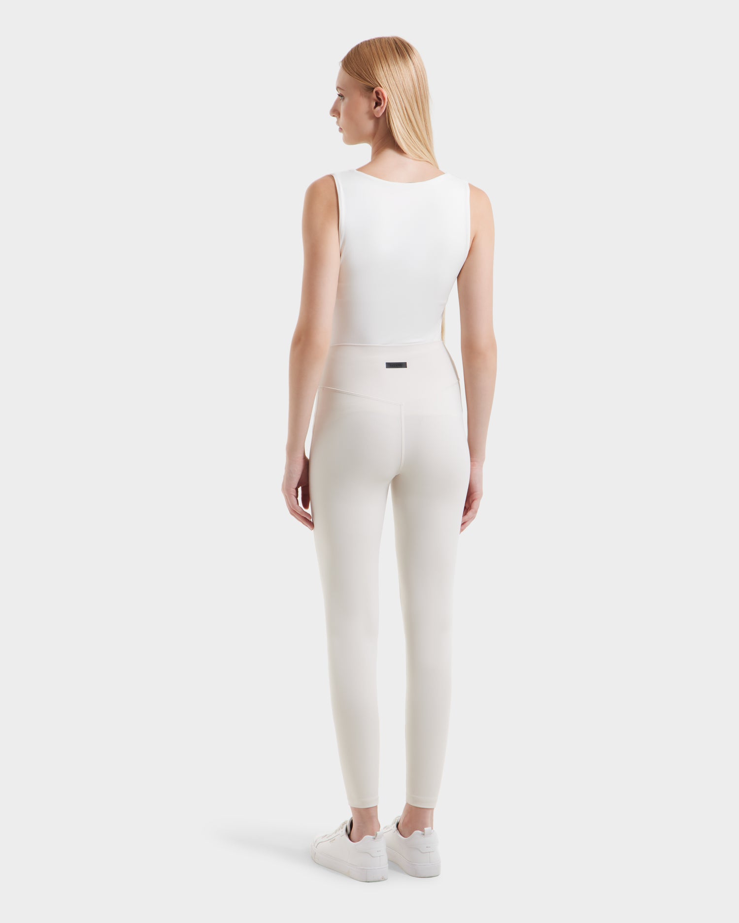 A woman with straight blonde hair wears a sleeveless white top, white sneakers, and high-waisted Rudsak SONOMA - 8525071 Vanilla leggings, standing facing away from the camera against a plain light background.