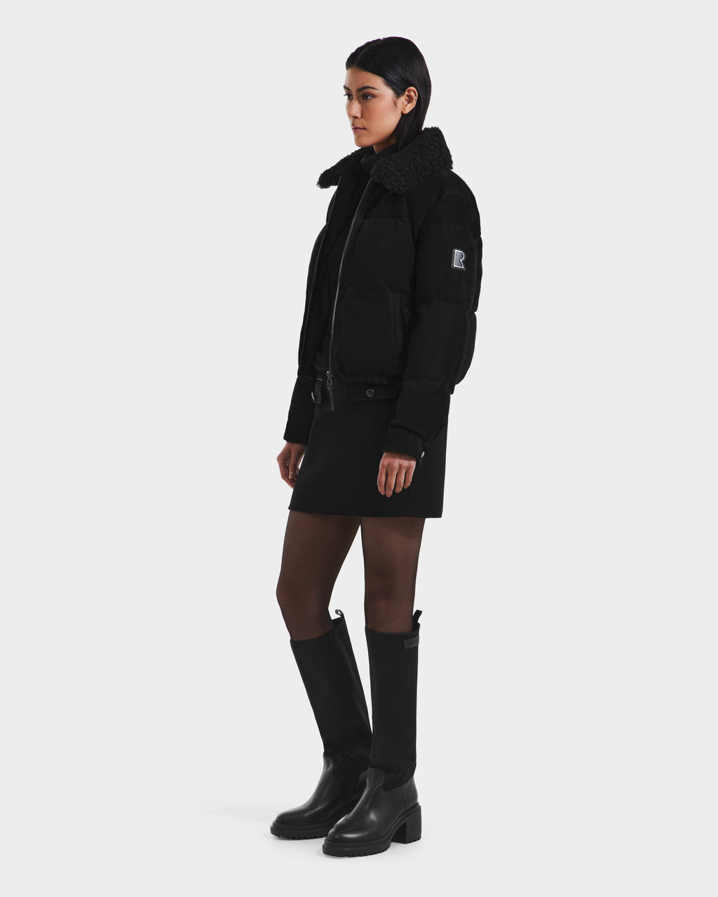 A woman with short dark hair and a neutral expression models the Rudsak STACEY - 8125049 Black shearling collar puffer jacket, styled as a cropped aviator, paired with a black mini skirt, knee-high boots, and dark tights against a plain background.
