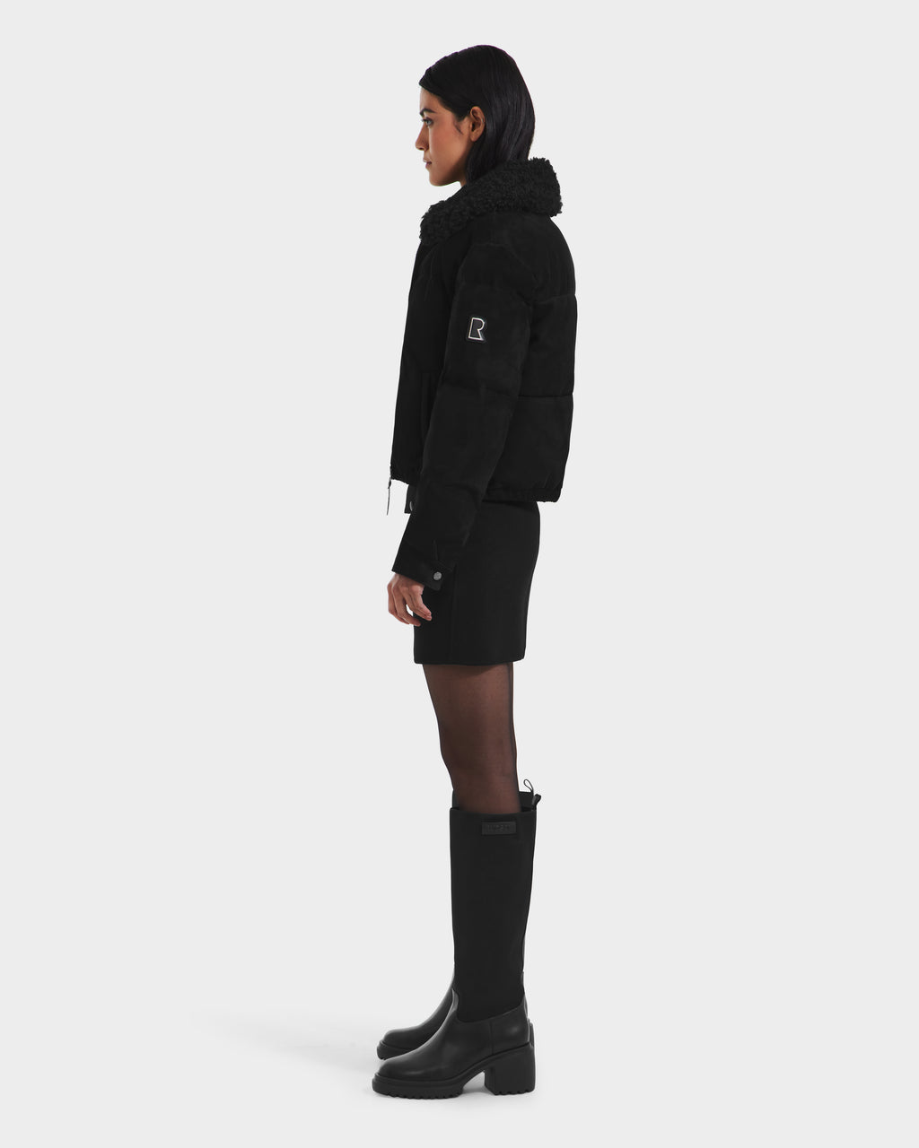 A person with straight dark hair tucked behind their ear stands in profile against a white background, wearing the Rudsak STACEY - 8125049 Black women’s cropped aviator jacket with a shearling collar, black skirt, and tall boots.