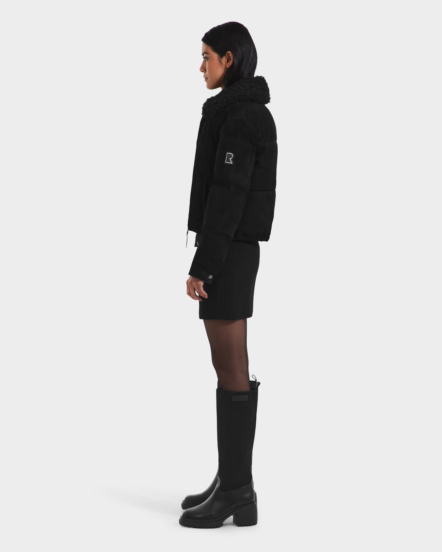 A person with straight dark hair tucked behind their ear stands in profile against a white background, wearing the Rudsak STACEY - 8125049 Black women’s cropped aviator jacket with a shearling collar, black skirt, and tall boots.