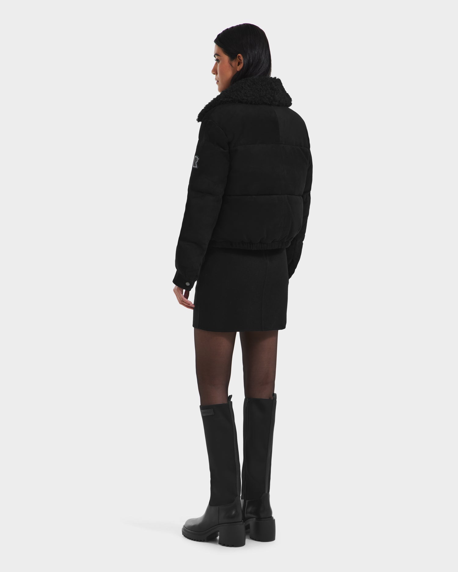 A woman faces away, wearing the Rudsak STACEY - 8125049 Black womens cropped aviator jacket with a shearling collar, a short black skirt, tights, and knee-high black boots against a plain white background.