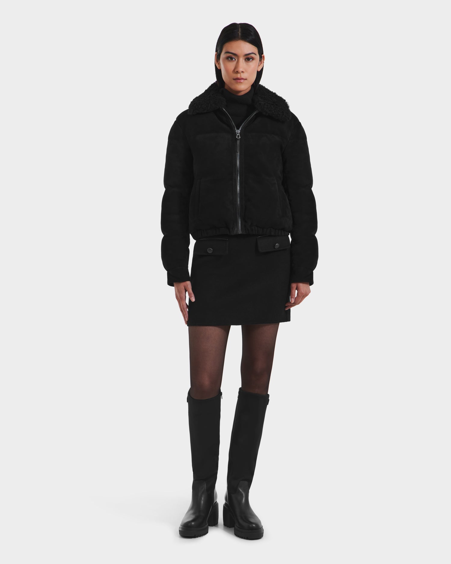 A woman stands against a plain background wearing the Rudsak STACEY - 8125049 Black cropped aviator jacket with a shearling collar, paired with a black turtleneck, mini skirt, sheer tights, and knee-high boots. She looks ahead with a neutral expression.