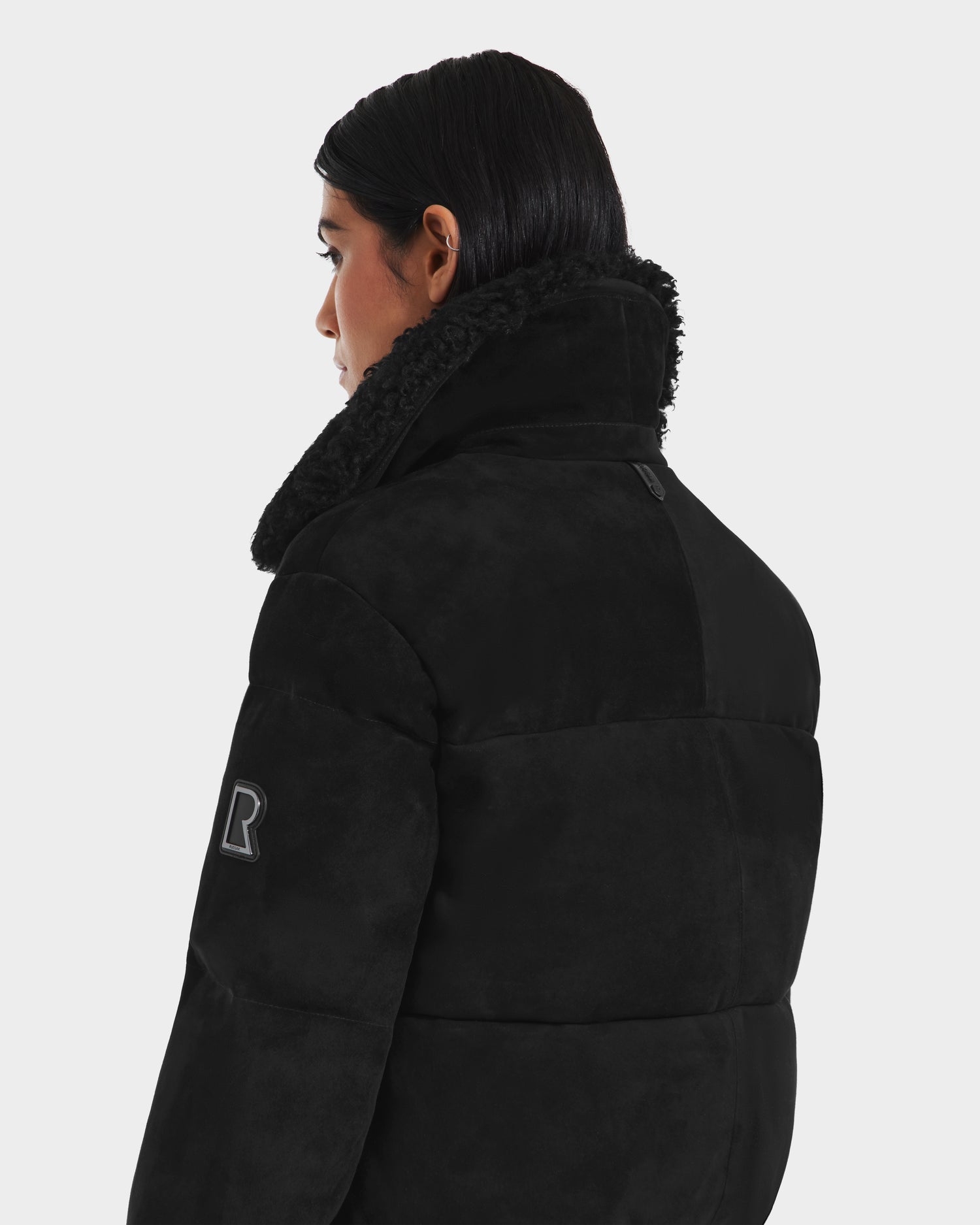 A person with dark hair models the Rudsak STACEY - 8125049 Black womens cropped aviator jacket with a shearling collar and an R patch on the sleeve, shown from the back and side against a plain light background.