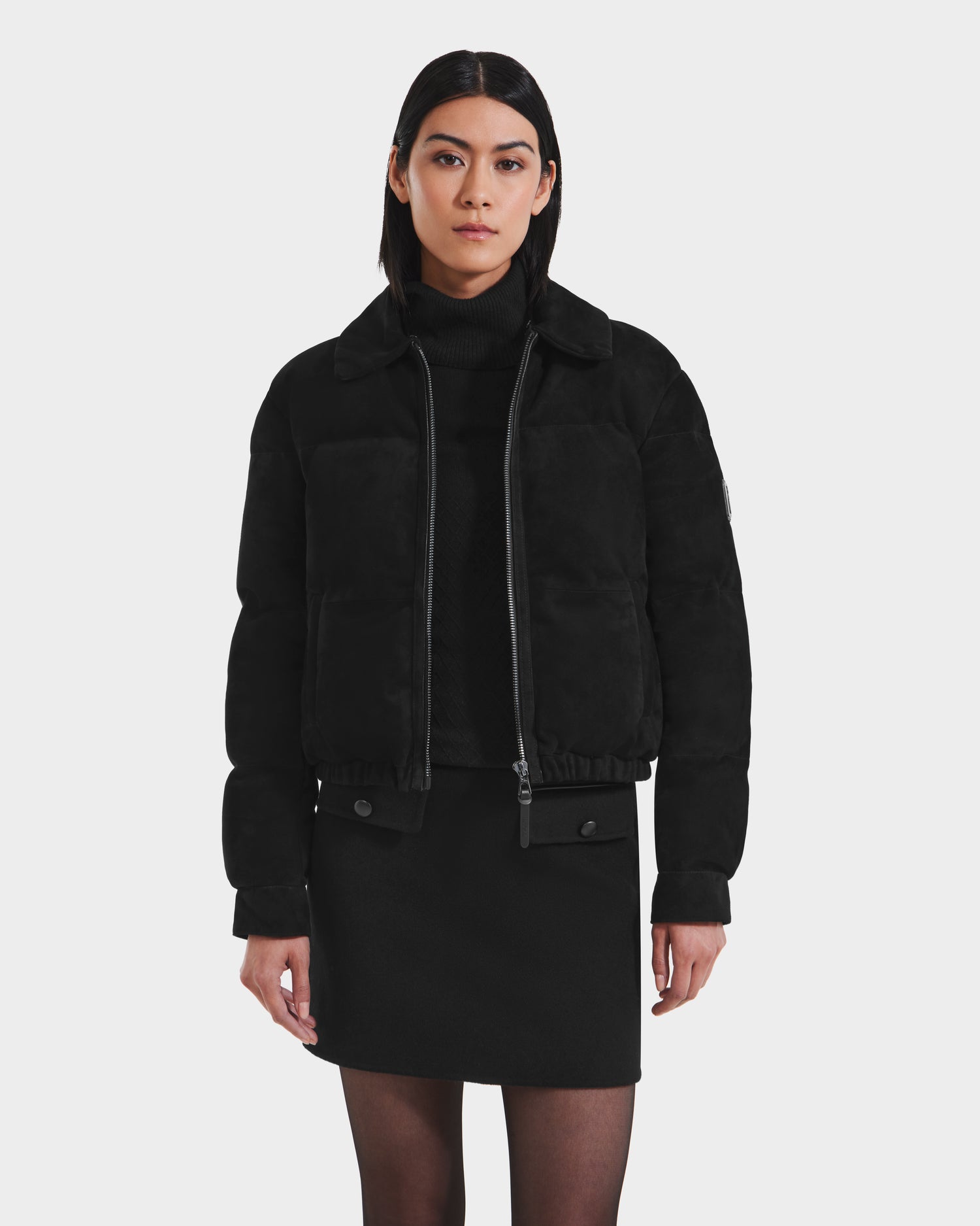 A woman with straight dark hair models the Rudsak STACEY - 8125049 Black women’s cropped aviator jacket with a shearling collar over a black turtleneck and skirt, standing against a plain white background.