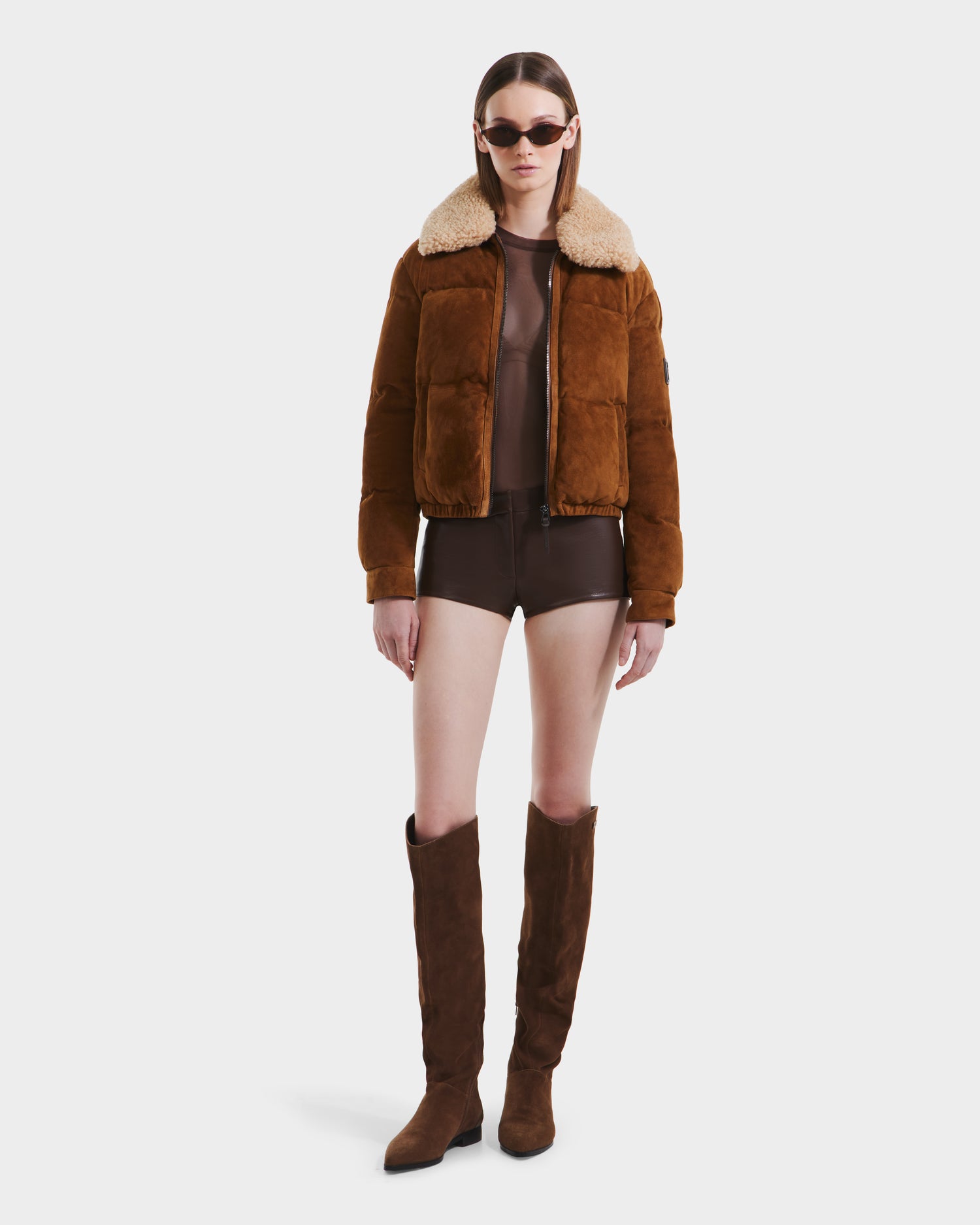 A person wearing the Rudsak STACEY - 8125049 Mocha women’s cropped bomber jacket with a shearling collar, styled with brown shorts, tall suede boots, and sunglasses stands against a plain white background.