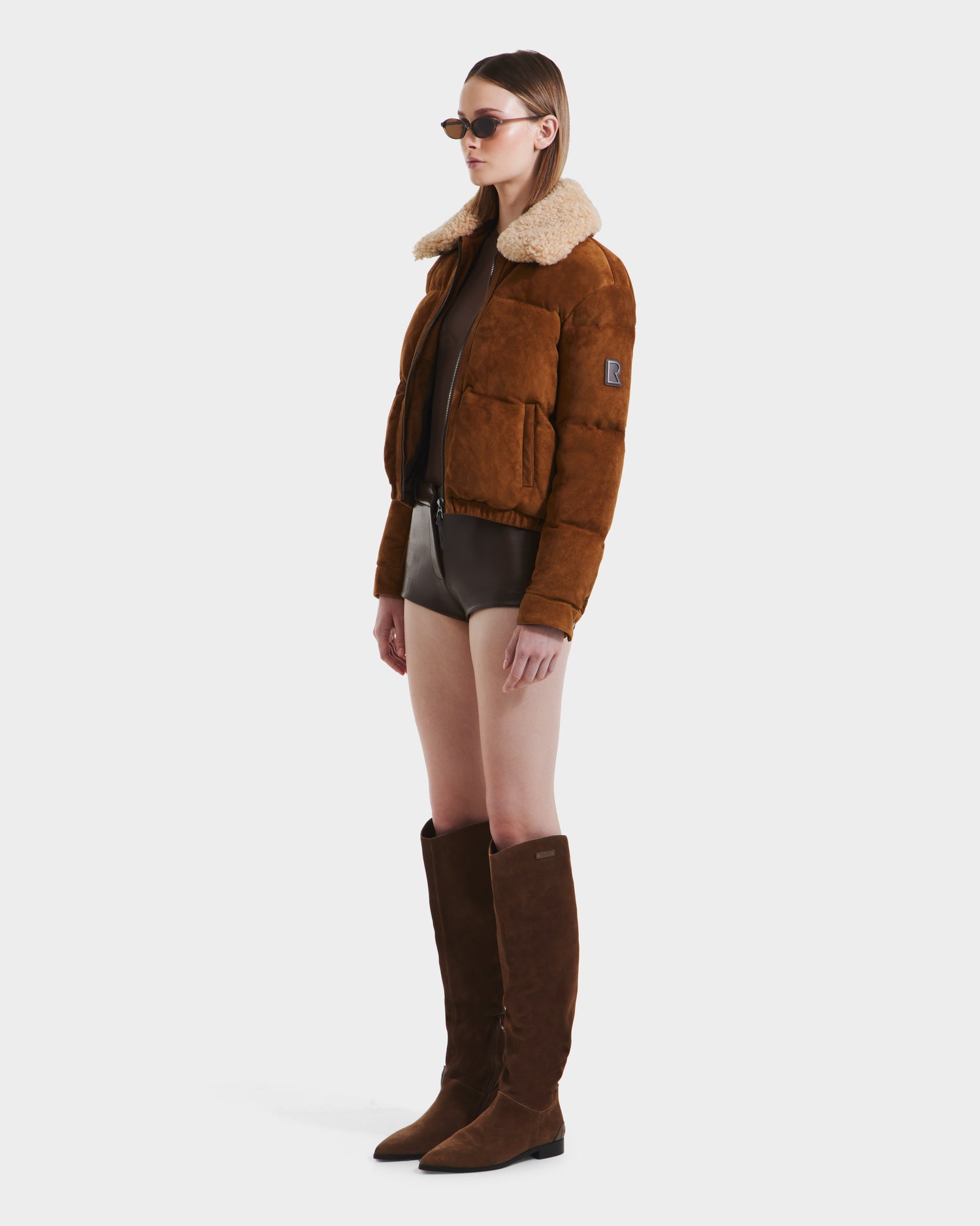 A woman wearing the Rudsak STACEY - 8125049 Mocha suede aviator jacket with a shearling collar, sunglasses, a brown bodysuit, and tall brown boots stands against a plain white background.