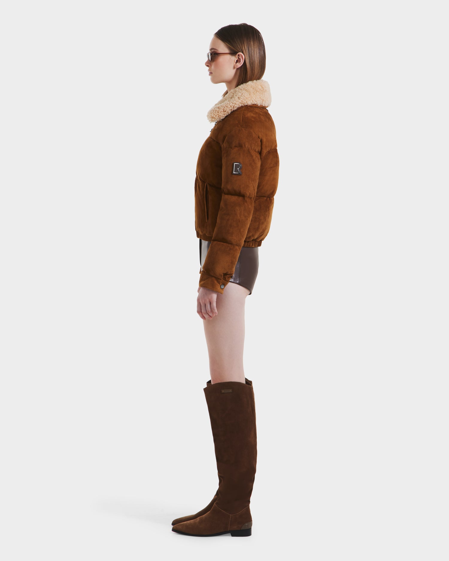A woman in profile wears Rudsaks STACEY - 8125049 Mocha womens cropped bomber jacket with a shearling collar, sunglasses, brown shorts, and knee-high brown suede boots against a white background.