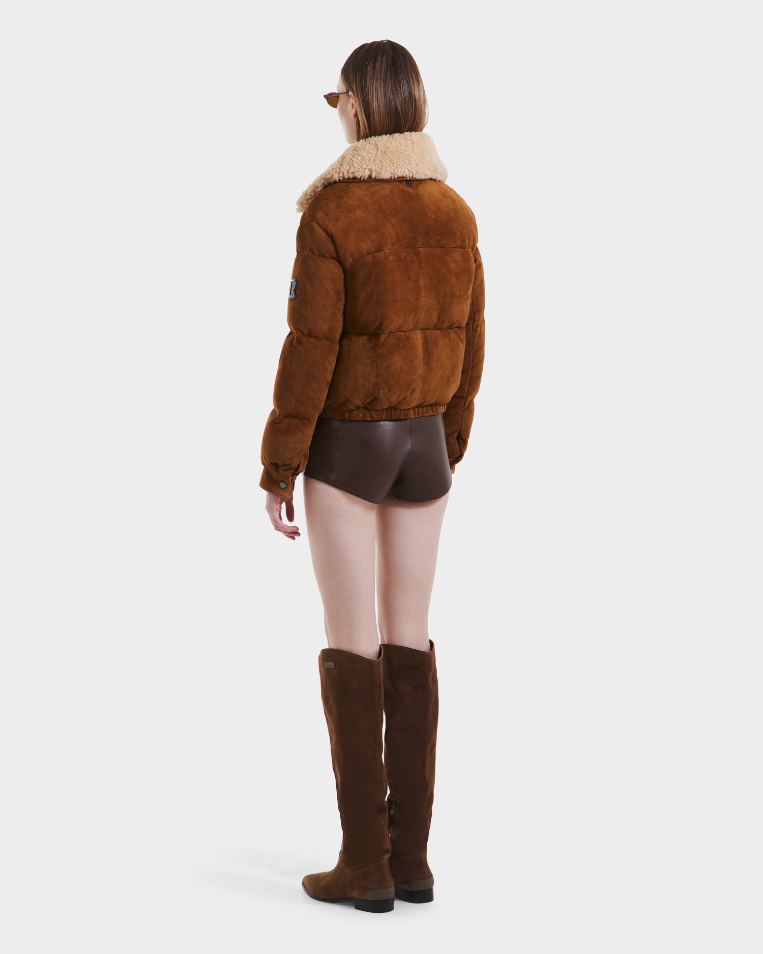 A person wearing Rudsaks STACEY - 8125049 Mocha womens cropped bomber jacket with a shearling collar, brown shorts, knee-high brown suede boots, and sunglasses stands facing away from the camera against a plain white background.