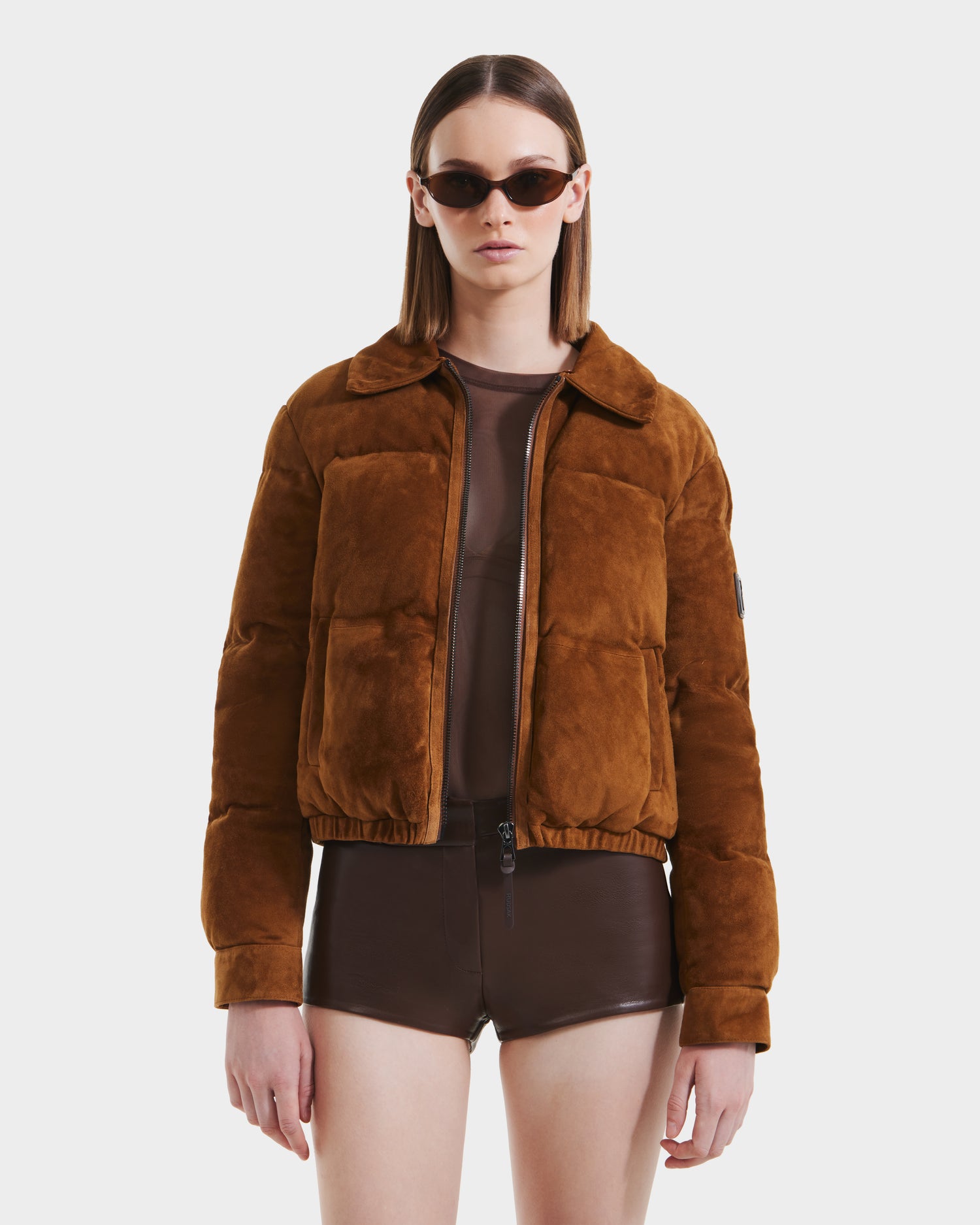 A person wearing the Rudsak STACEY - 8125049 Mocha cropped bomber jacket with a shearling collar, paired with dark sunglasses, a sheer brown top, and brown shorts, stands against a plain white background.
