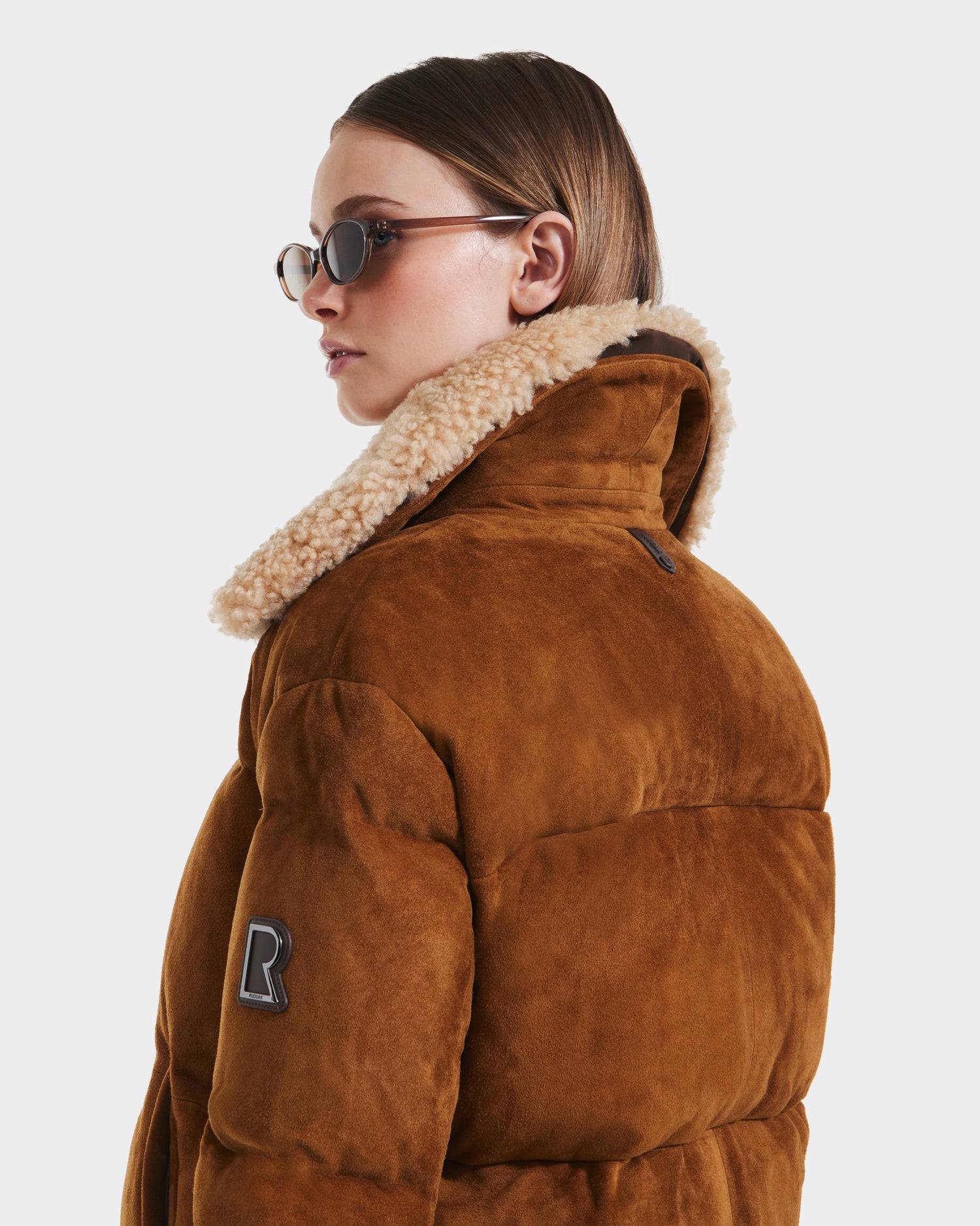 A person in oval sunglasses and the Rudsak STACEY - 8125049 Mocha women’s cropped bomber jacket with shearling collar, sporting a black R patch on the sleeve, looks over their shoulder against a white background.