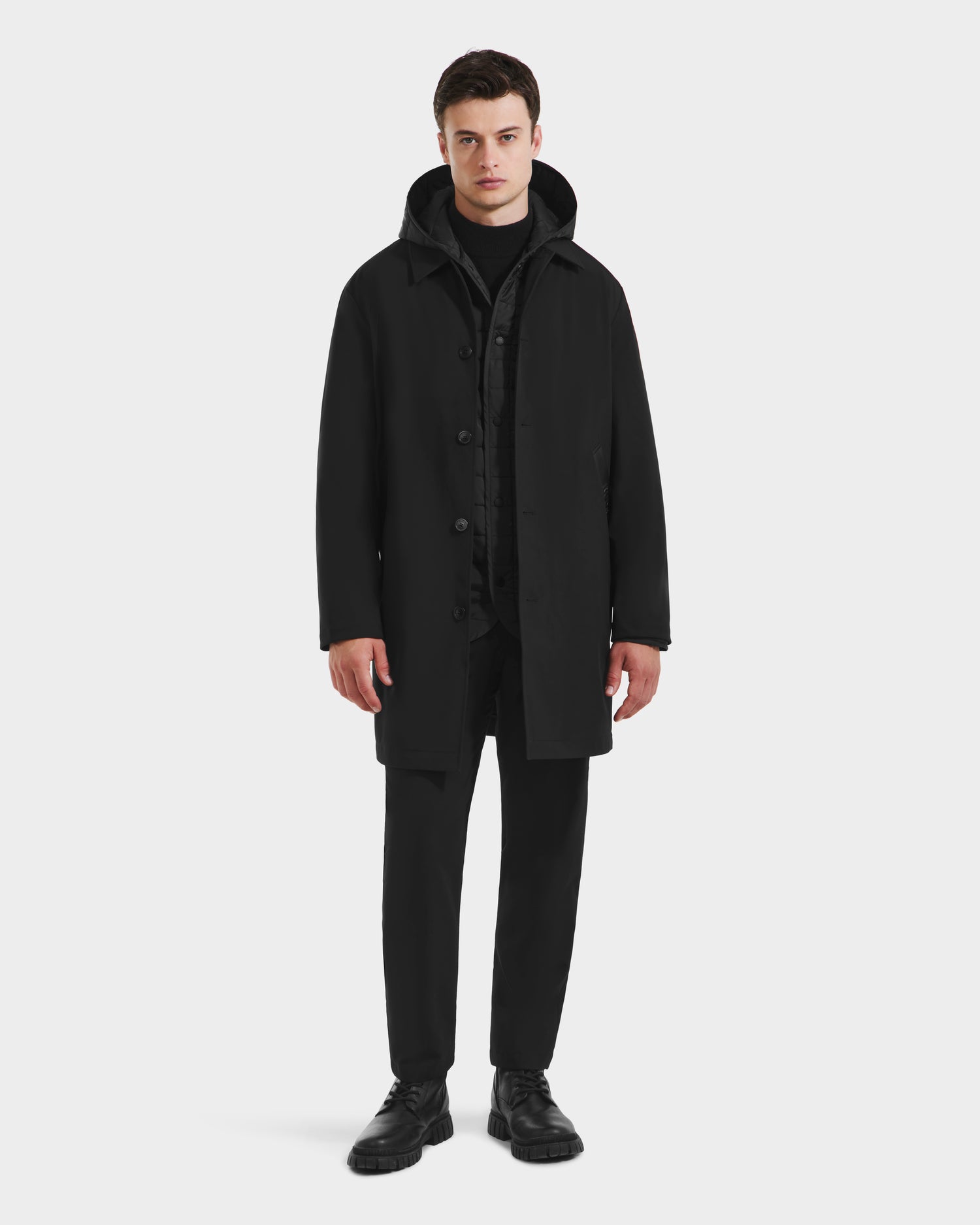 A man stands against a plain white background, wearing the Rudsak STANTON - 6125531 Black men’s 3-in-1 overcoat over a black outfit and black shoes. He faces forward with a neutral expression.