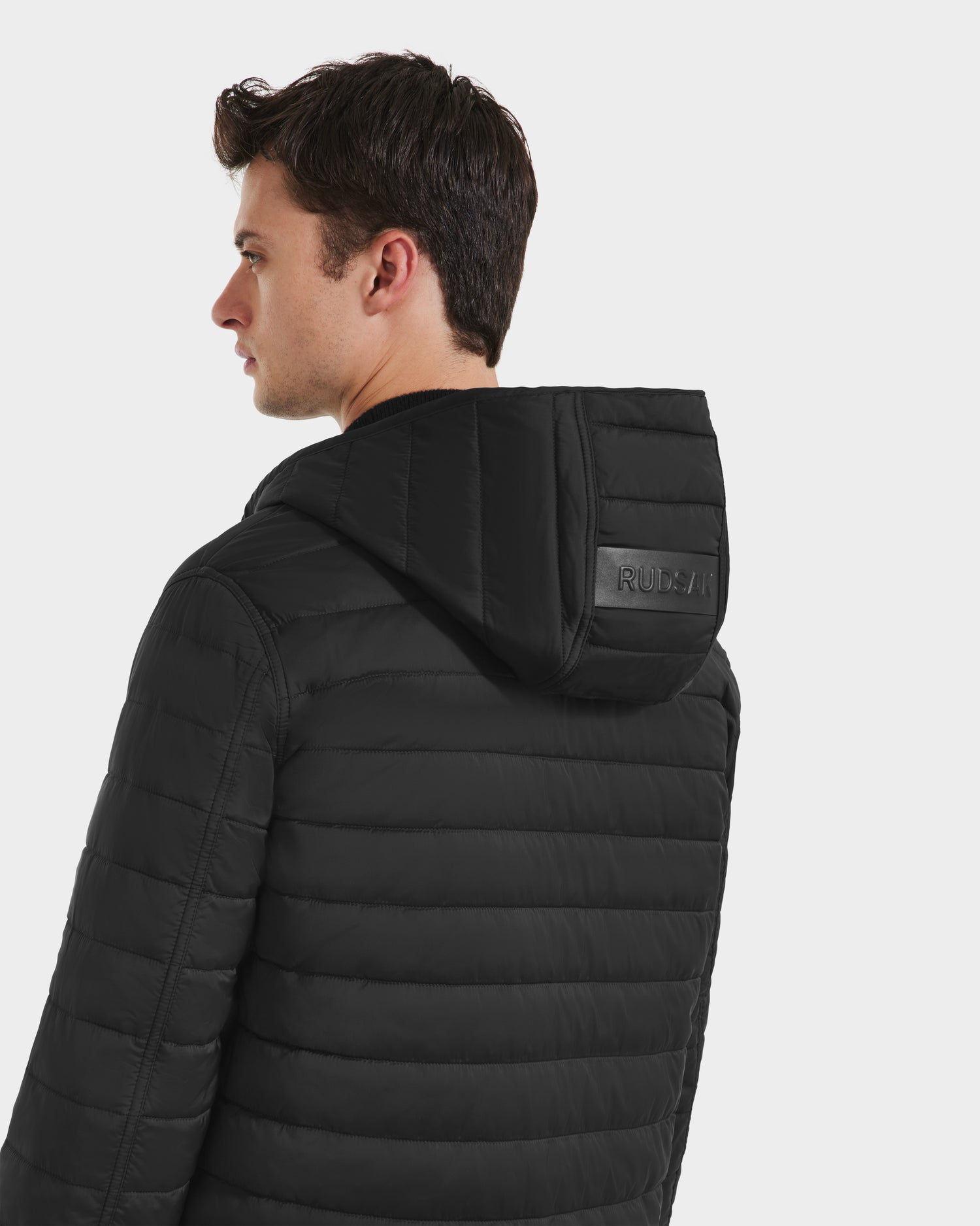 A young man with short brown hair faces away from the camera, wearing the Rudsak STANTON - 6125531 Black quilted puffer jacket with a hood and subtle logo detail, set against a plain white background.