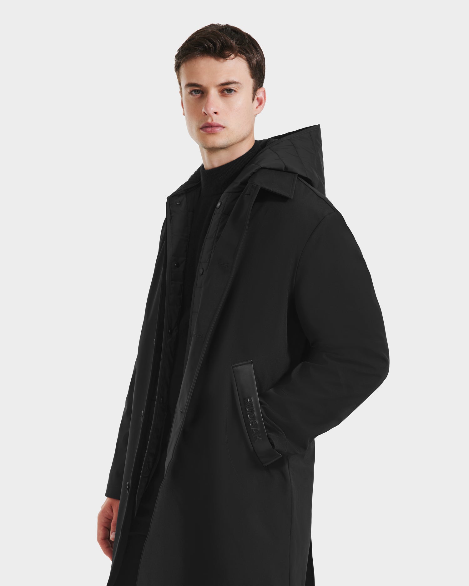 A young man with short brown hair wears the Rudsak STANTON - 6125531 Black men’s 3-in-1 overcoat, featuring a high collar and hood, with the technical shell layered over a quilted puffer, standing against a white background.