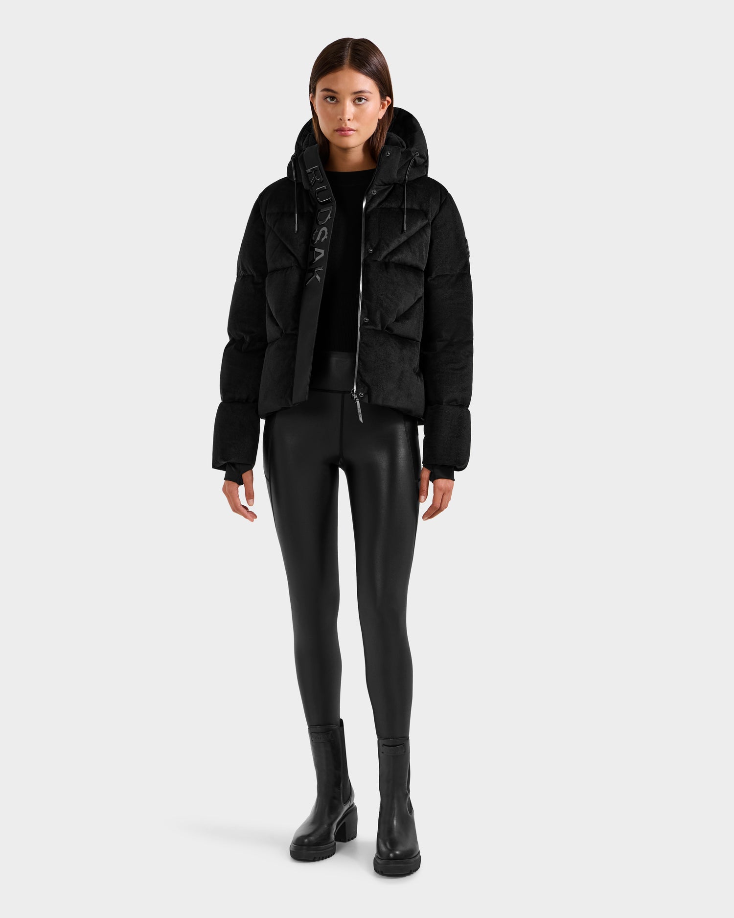A woman stands facing forward, wearing the Rudsak STEFANIE V - L125584 Black down puffer jacket with a hood, paired with black fitted leggings, a black top, and black ankle boots against a plain light gray background.