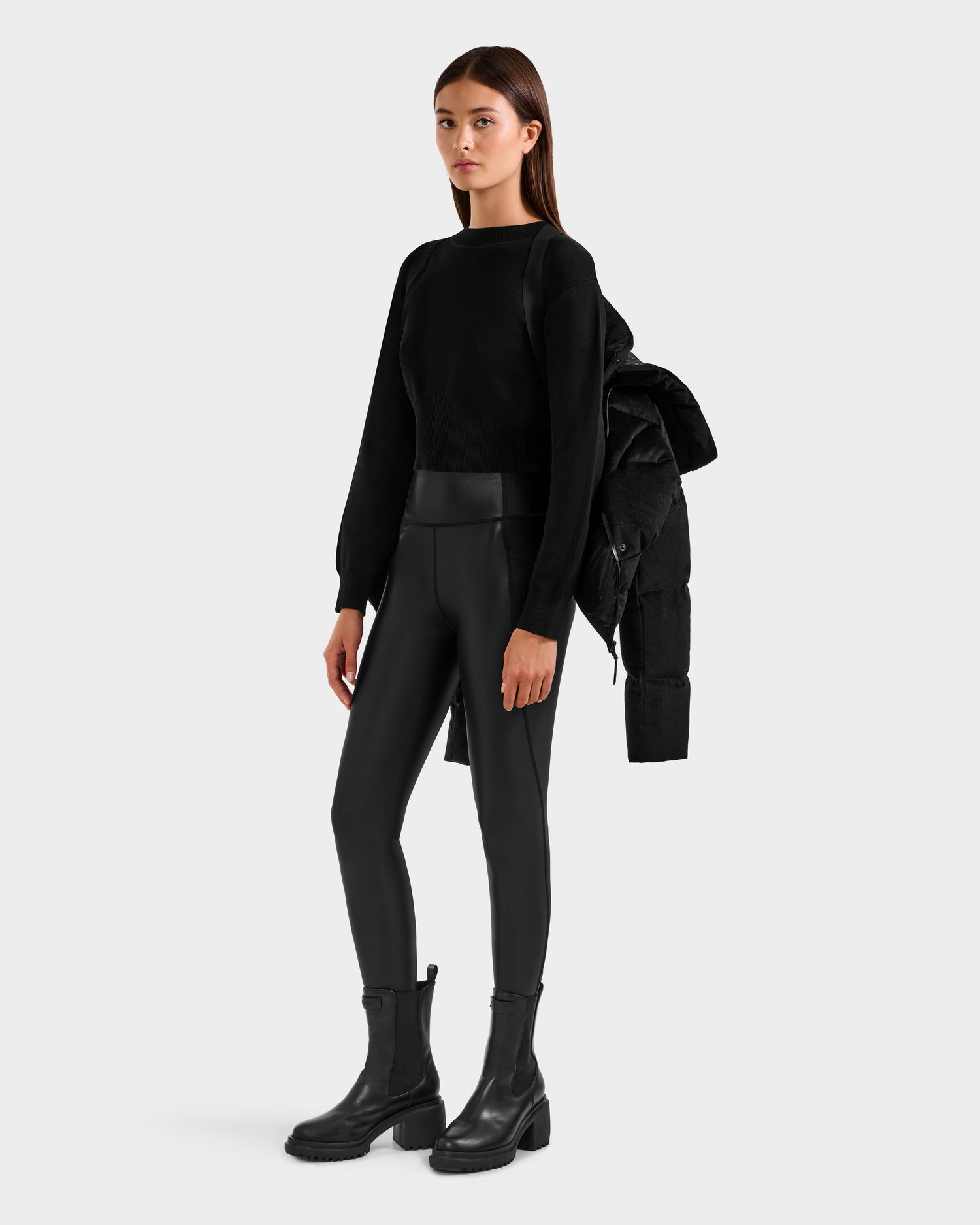 A woman poses against a plain background in a black long-sleeve top, black leather-look leggings, chunky boots, and the Rudsak STEFANIE V - L125584 Black down puffer jacket draped over one shoulder for a chic winter look.
