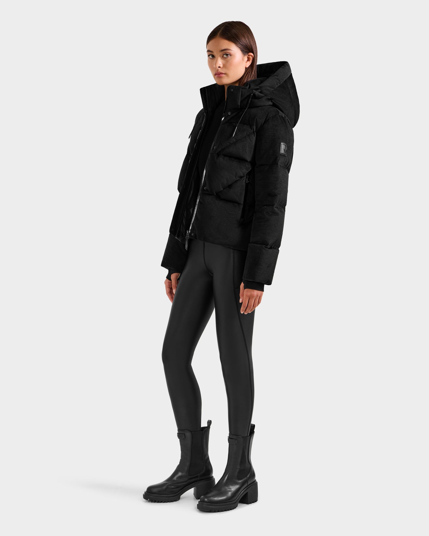 A woman models the Rudsak STEFANIE V - L125584 Black down puffer jacket, paired with black leggings and ankle boots, standing slightly angled against a plain light gray background.