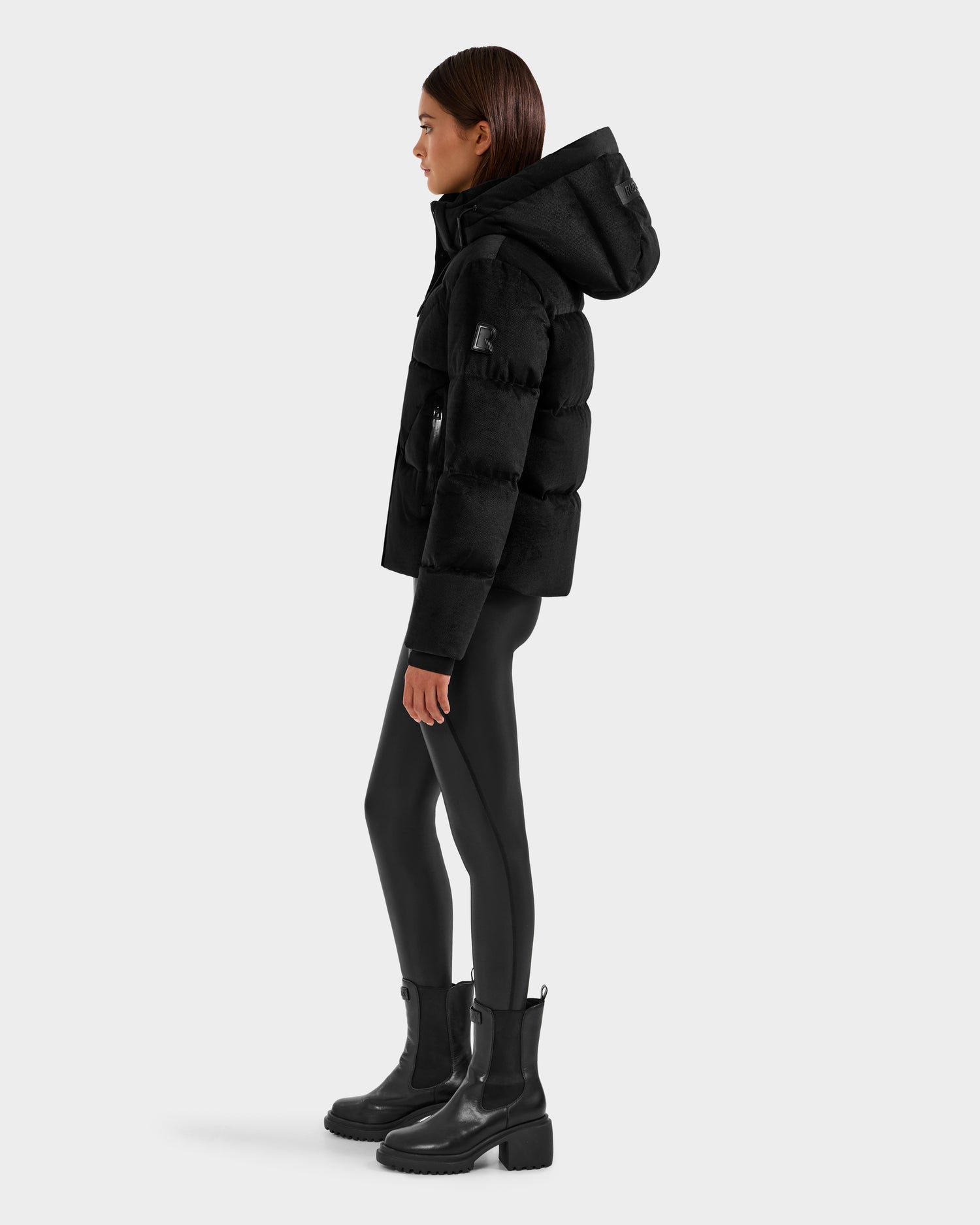 A woman stands in profile against a plain background, wearing the Rudsak STEFANIE V - L125584 Black down puffer with a hood, paired with black leggings and black ankle boots.