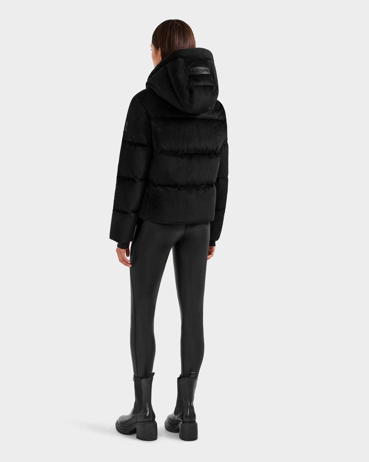 A person stands facing away against a light gray background, wearing the Rudsak STEFANIE V - L125584 Black down puffer jacket with a hood, paired with black shiny leggings and chunky boots.