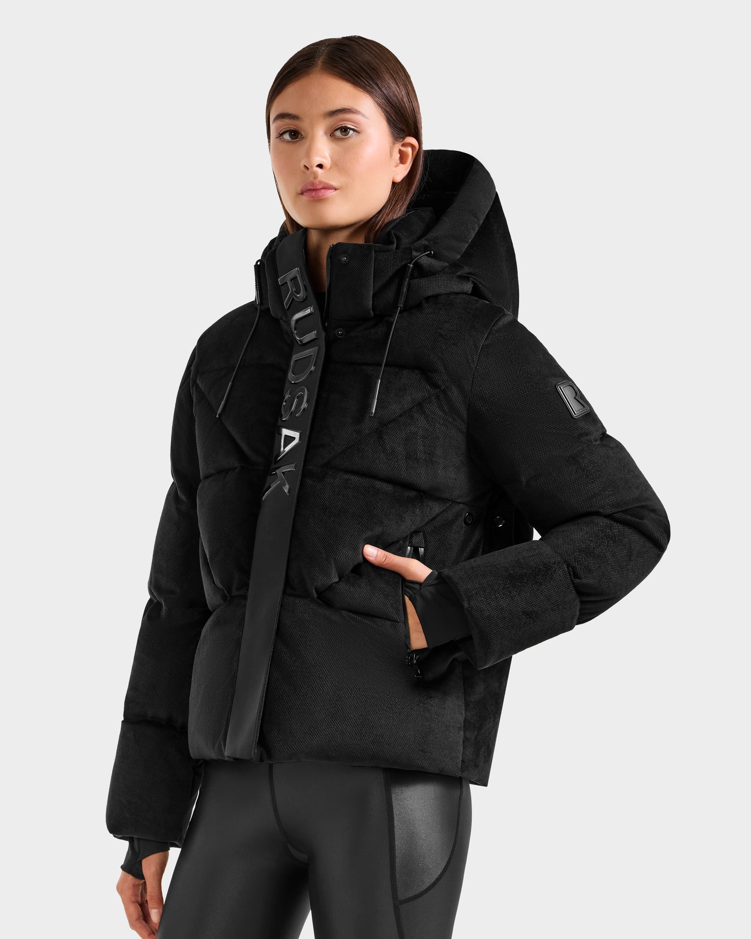 A woman stands confidently against a light gray background wearing the Rudsak STEFANIE V - L125584 black down puffer jacket with "RUDSAK" on the collar and black leggings—an essential piece for any winter wardrobe.