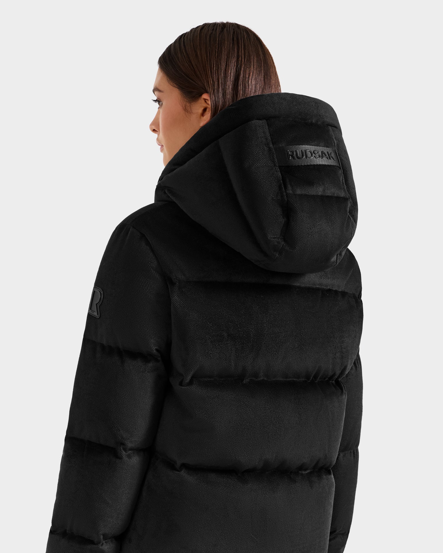 A woman with straight brown hair is wearing the Rudsak STEFANIE V - L125584 black down puffer jacket, featuring a hood with the brand name on the back and a sleeve patch. She’s facing away and slightly to the right.