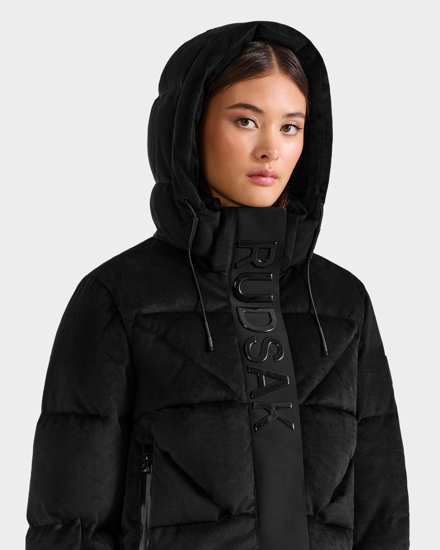 A person wears the Rudsak STEFANIE V - L125584 Black down puffer jacket, featuring bold Rudsak branding across the front panel. The background is plain light gray.