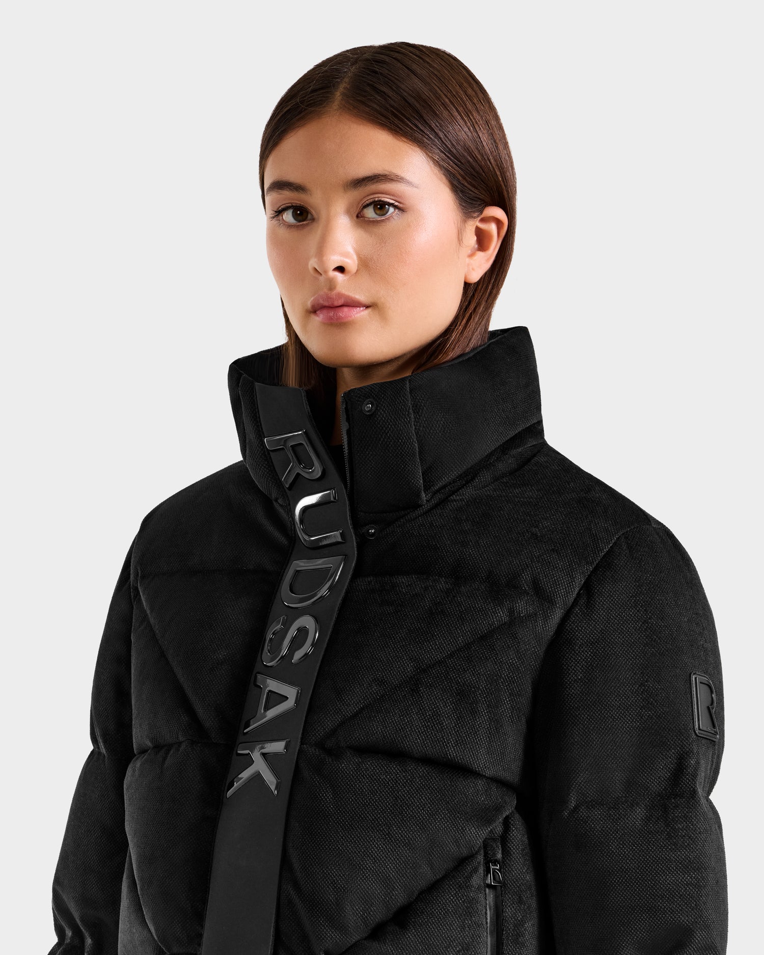 A young woman with straight brown hair wears the Rudsak STEFANIE V - L125584 black puffer jacket with a high collar featuring RUDSAK lettering and a logo patch on the sleeve, standing against a light background.