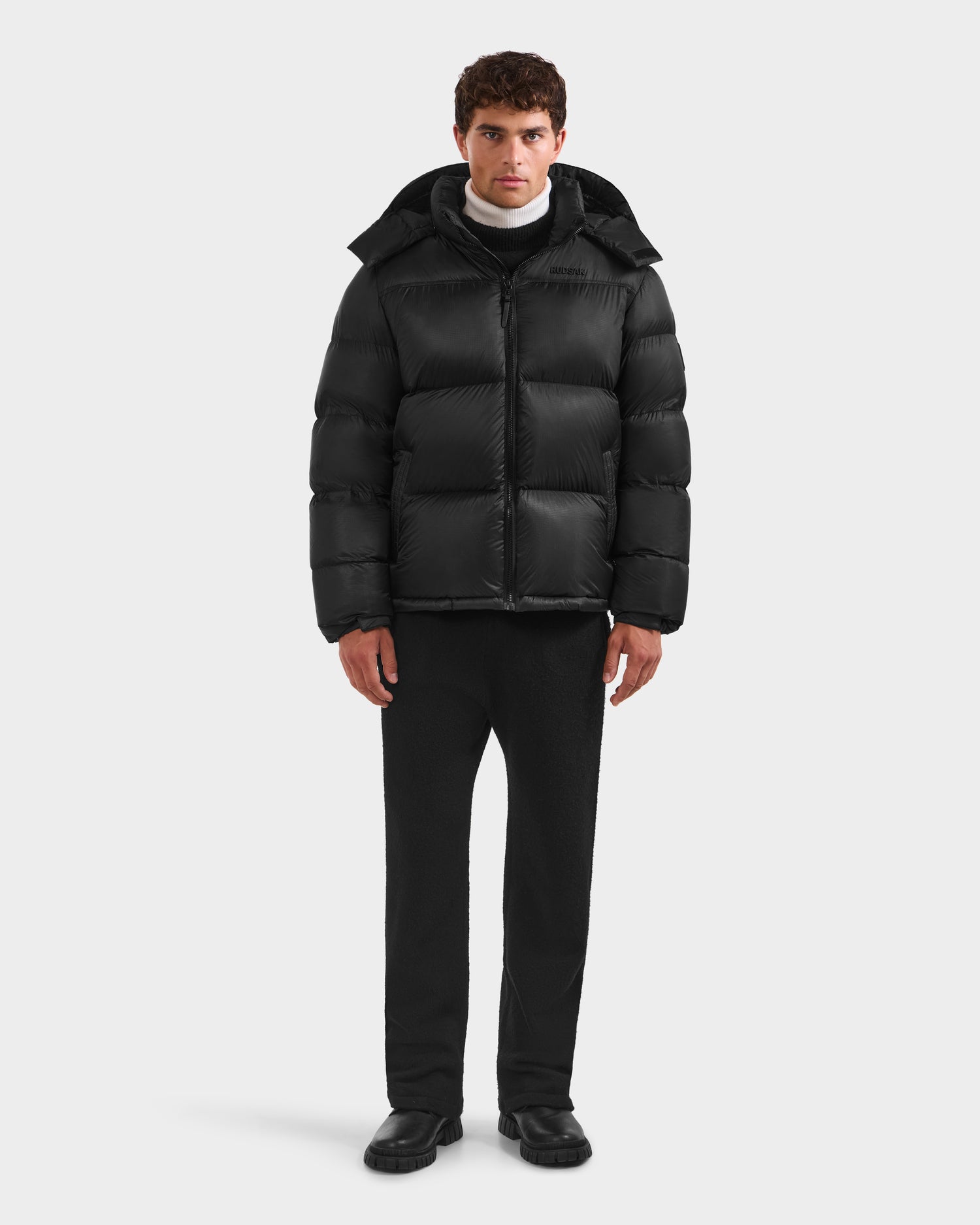 A man faces forward against a light gray background, wearing the Rudsak STEFANO - 6125574 Black men’s down puffer jacket with a hood, paired with black pants, black shoes, and a white turtleneck.
