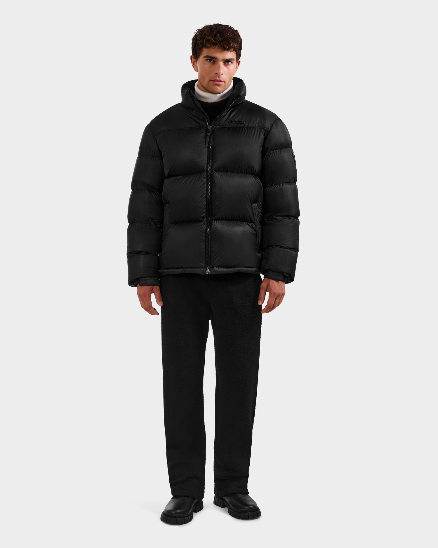 A man stands facing forward in the Rudsak STEFANO - 6125574 Black men's down puffer jacket, with black pants and shoes, against a plain light gray background.