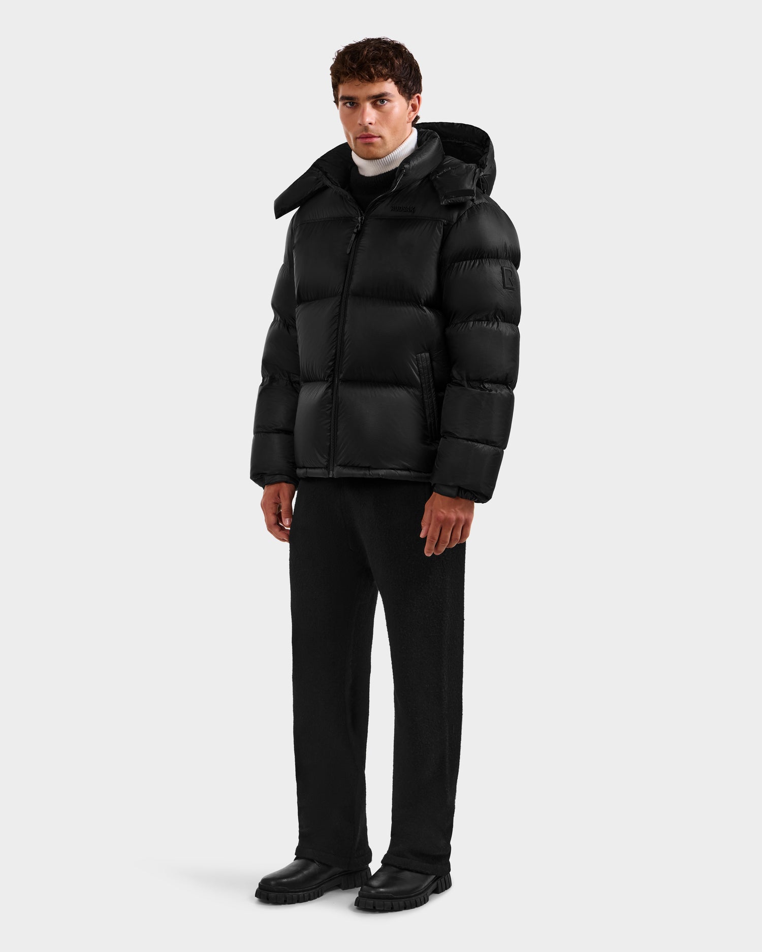 A man with short, dark hair wears the Rudsak STEFANO - 6125574 Black men’s down puffer over a white turtleneck, paired with black pants and shoes. The background is plain and light gray.