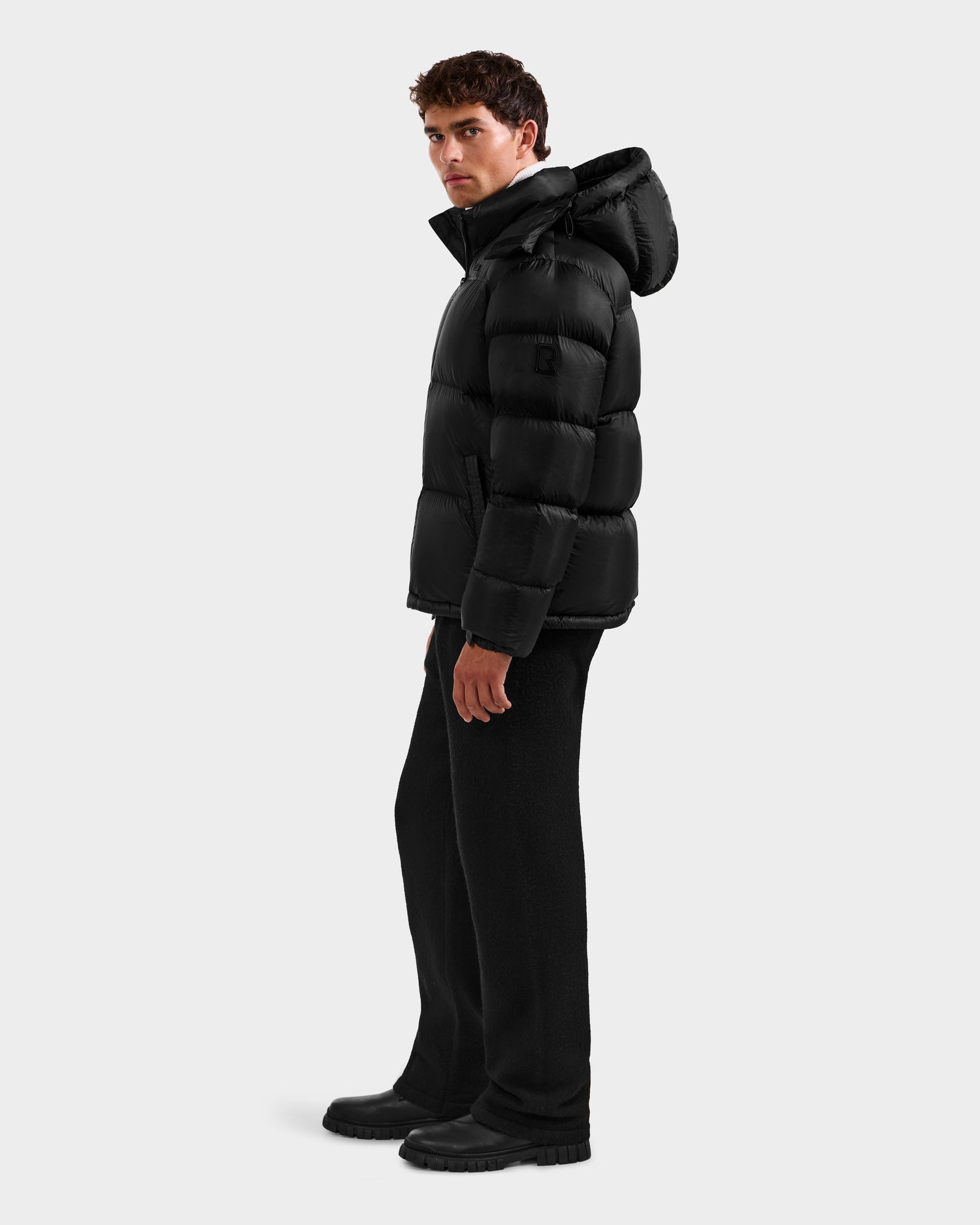 A man stands in profile wearing the Rudsak STEFANO - 6125574 Black men’s down puffer jacket, paired with black pants and boots, against a plain light gray background.