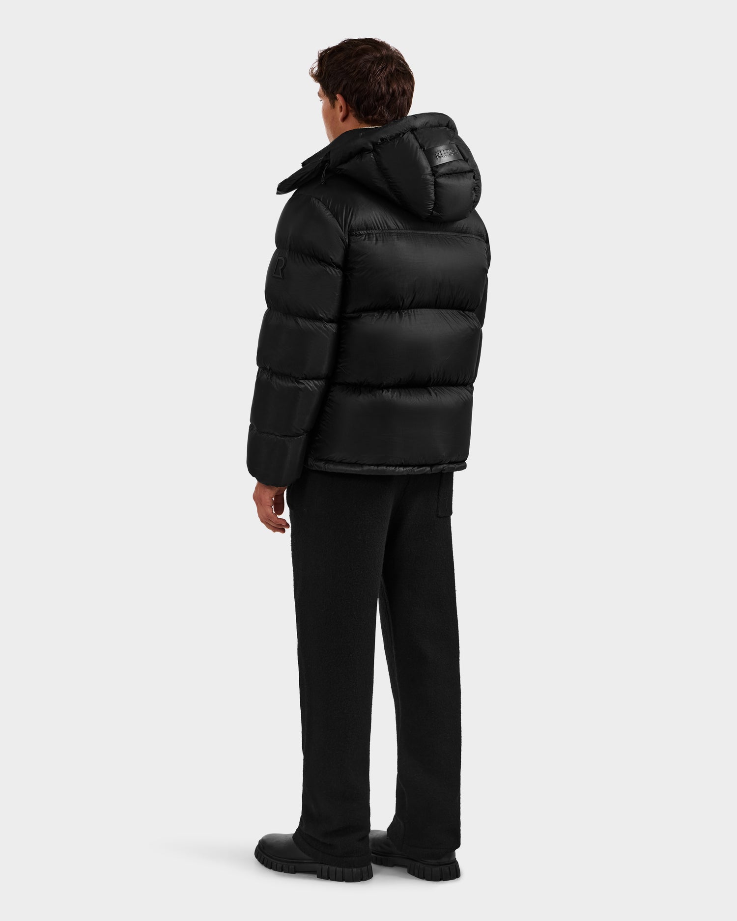 A man faces away, wearing Rudsak's STEFANO - 6125574 Black men's down puffer jacket with black pants and shoes, set against a plain, light backdrop.