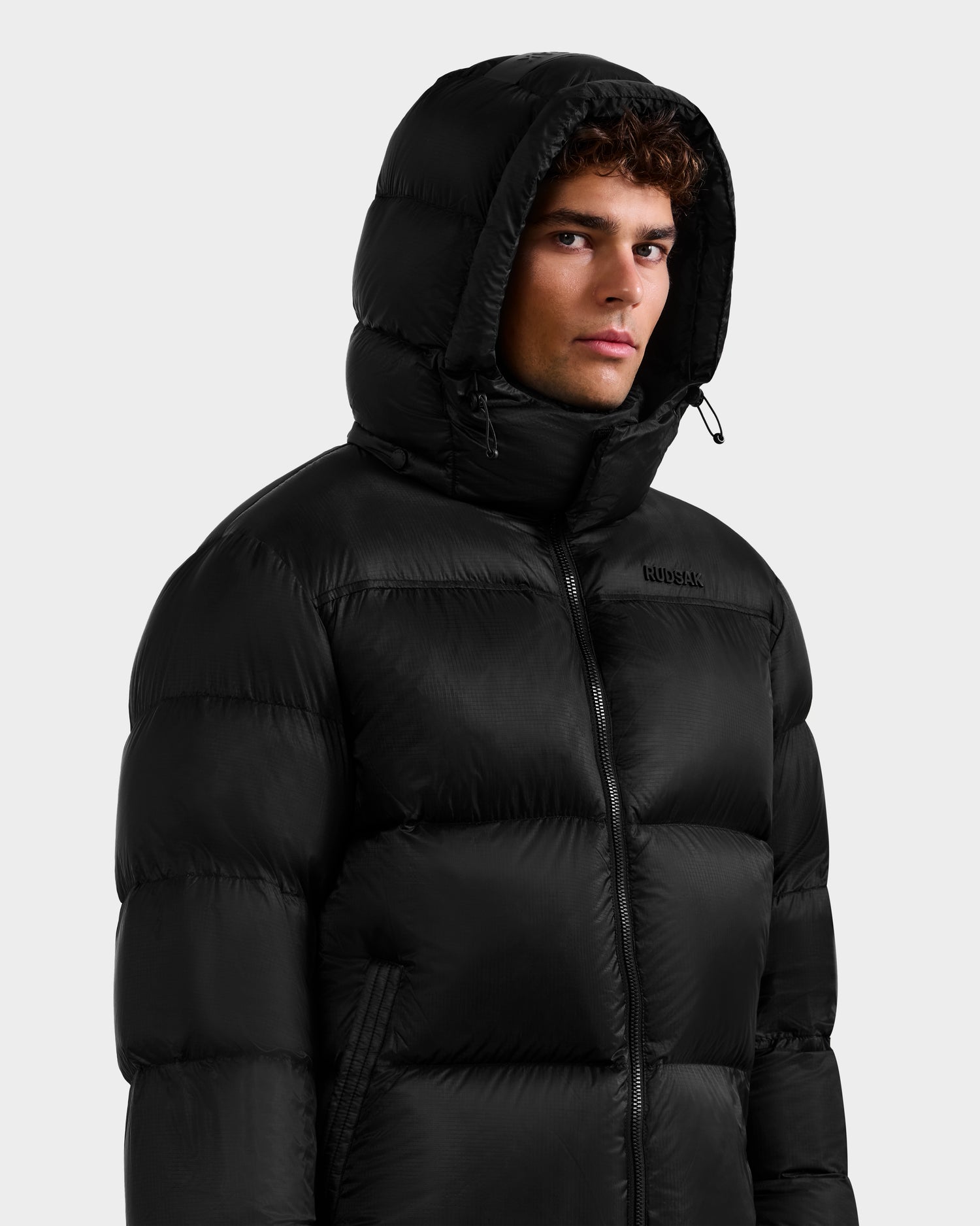A young man wears the Rudsak STEFANO - 6125574 Black men’s down puffer against a plain light background, gazing to the side with a neutral expression—a sleek and technical outerwear statement.