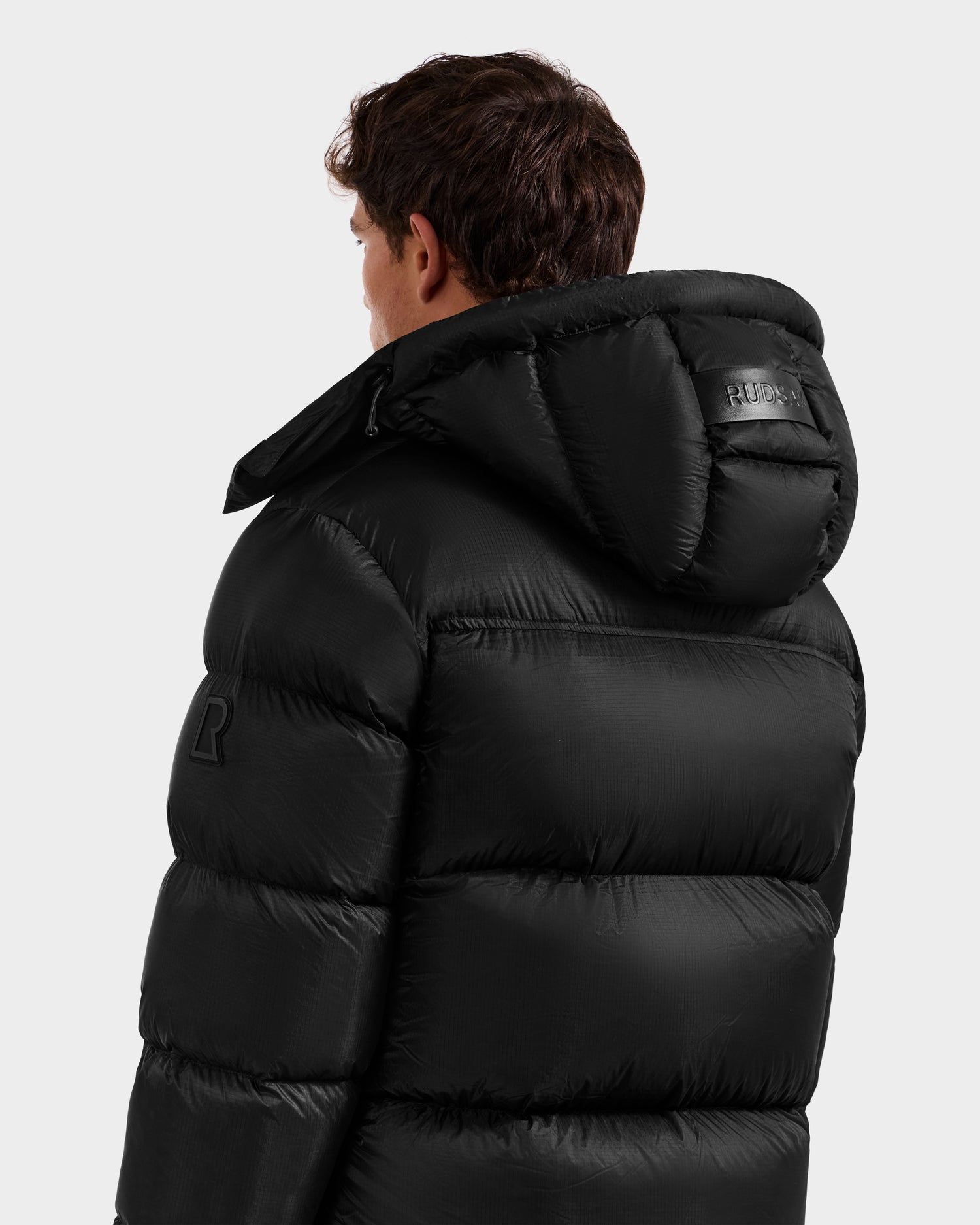 A person facing away wears the Rudsak STEFANO - 6125574 Black men’s down puffer jacket with a hood, visible stitching, and patch logos on the arm and hood. The technical outerwear stands out against a plain light gray background.