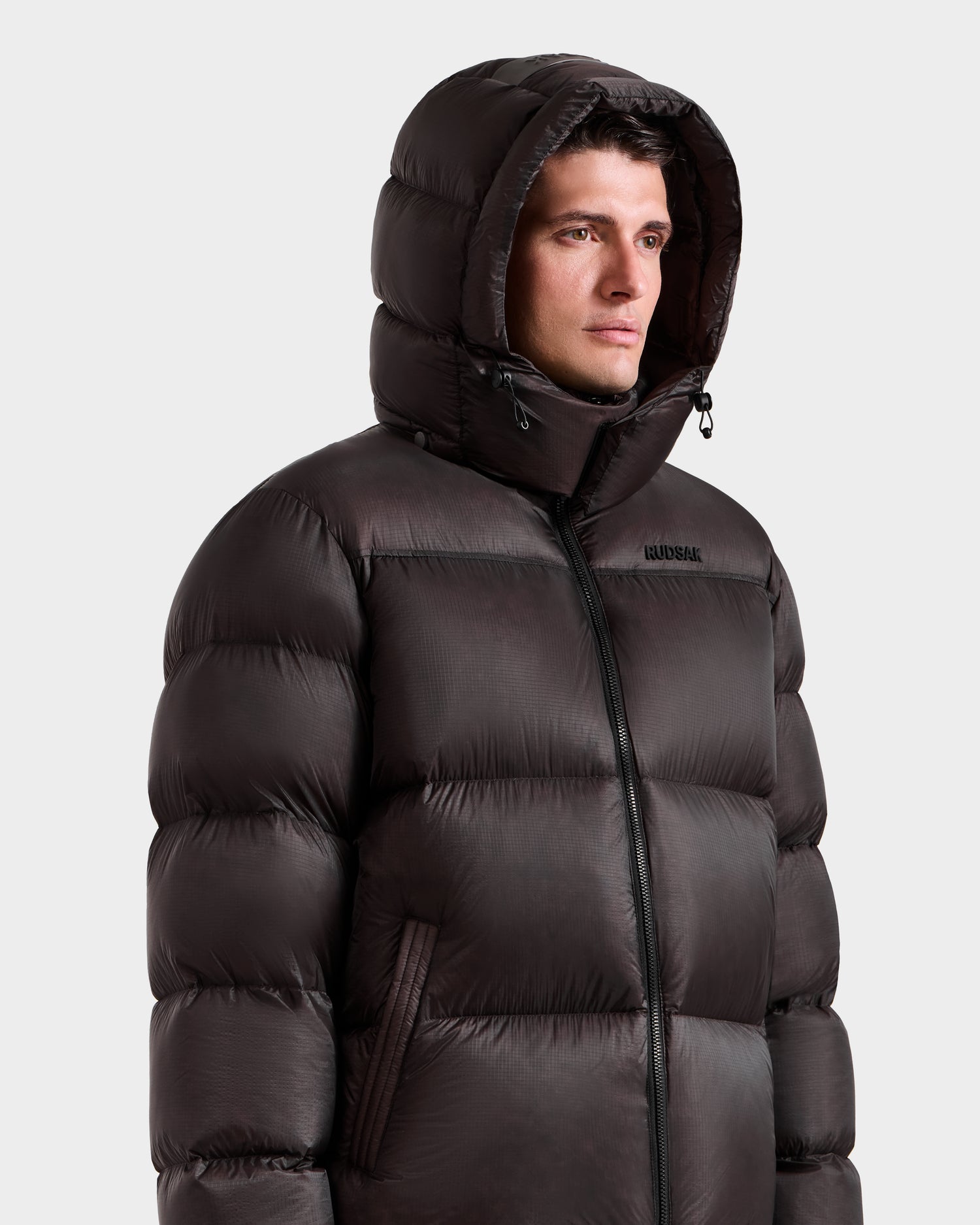 A man wears the Rudsak STEFANO - 6125574 Espresso down puffer jacket, featuring a hood and horizontal quilting, zipped up and facing slightly to the side against a plain light background.