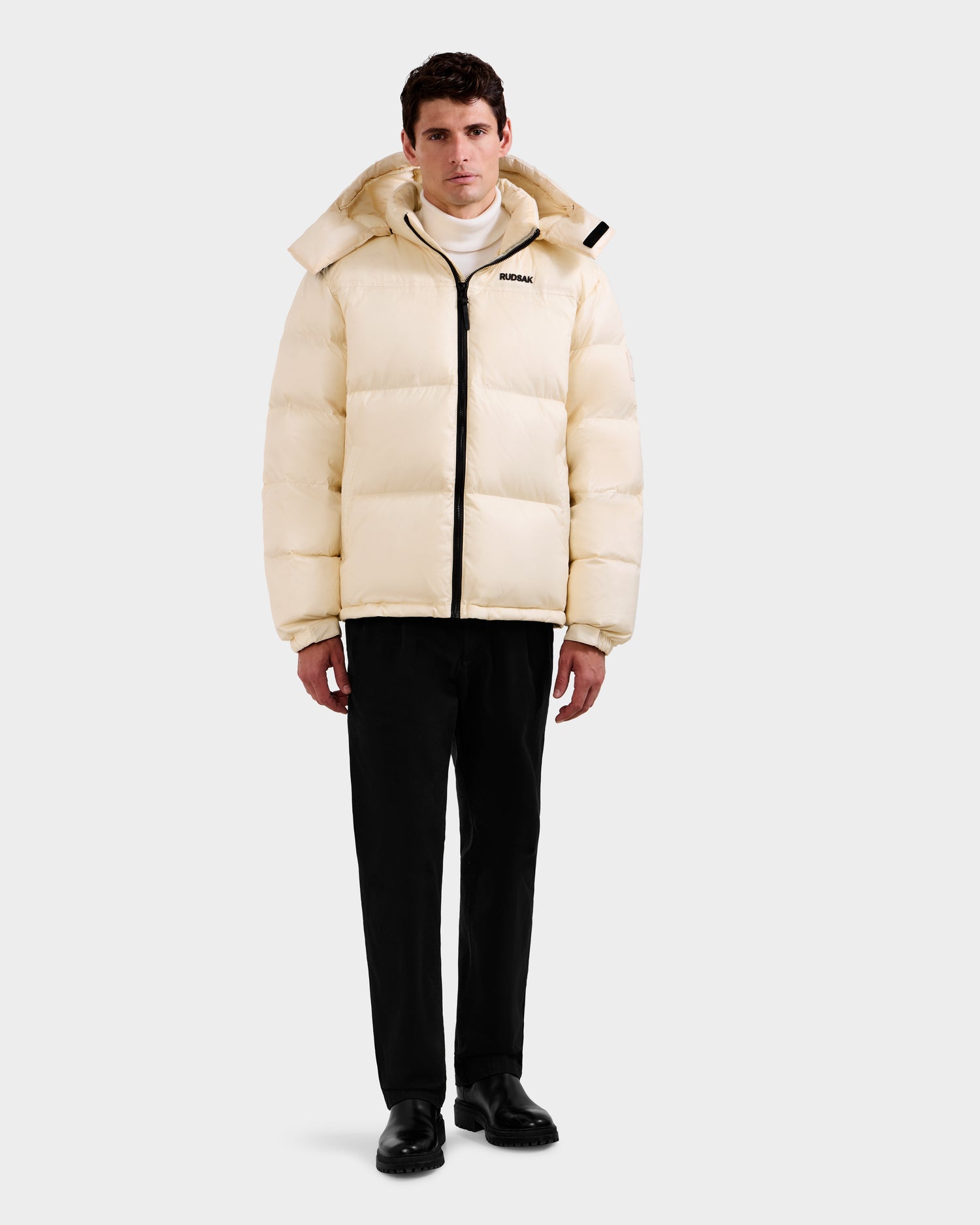 A man stands facing forward in the Rudsak STEFANO - 6125574 Vanilla men’s down puffer jacket with hood, black pants and shoes, and a white turtleneck. The RDS-certified down jacket features a black zipper and a small chest logo.