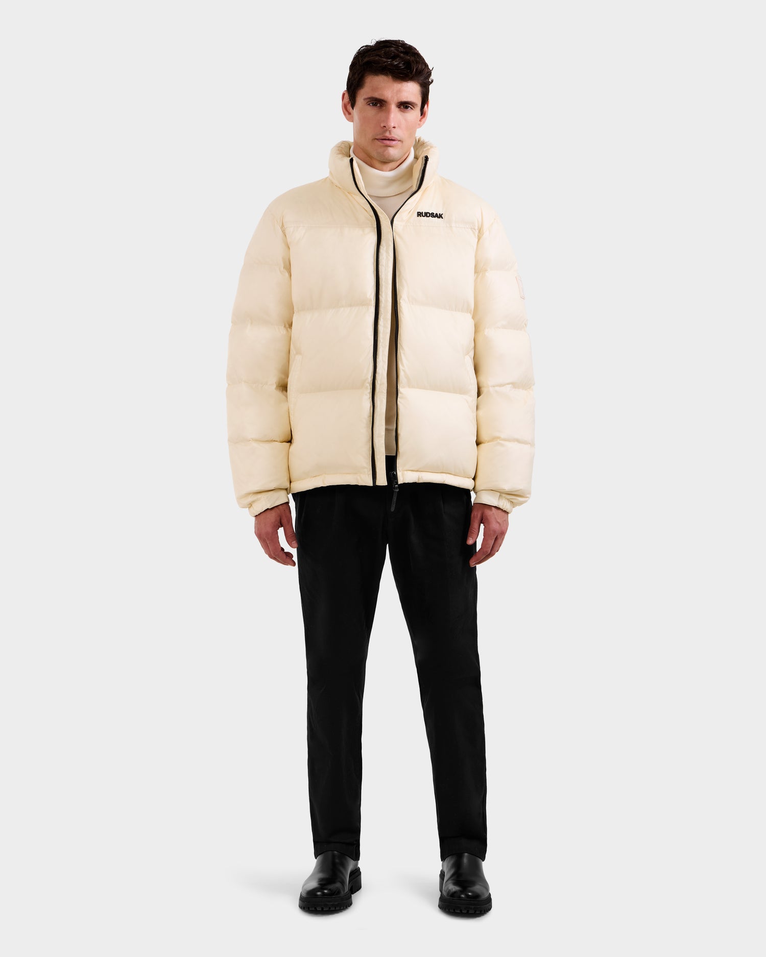 A man faces forward wearing the Rudsak STEFANO - 6125574 Vanilla men’s down puffer jacket with RDS-certified down, black pants, and black shoes against a plain light gray background.