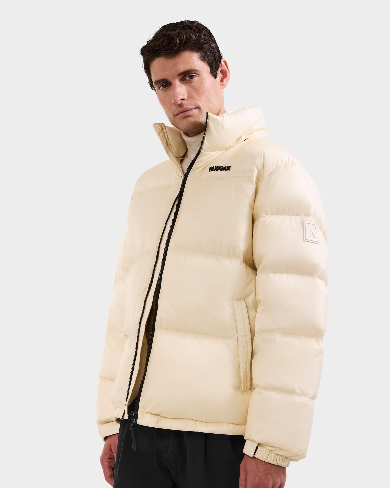 A man wears the STEFANO - 6125574 Vanilla men’s down puffer jacket by Rudsak, featuring a high collar, black zipper, "RUPSAR" on the chest, and a subtle logo on the sleeve, against a plain light background.